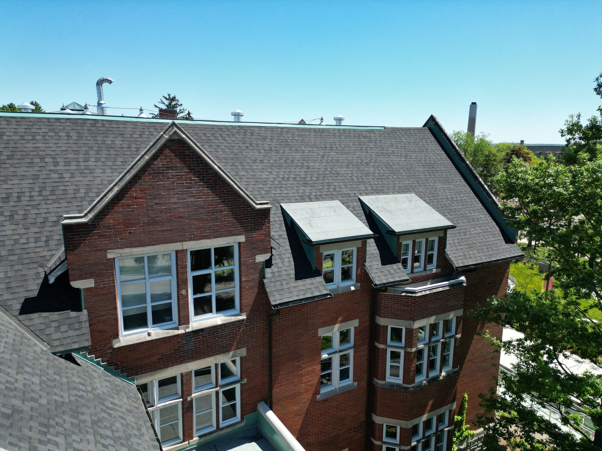 A multi-story brick building with a dark shingled roof, featuring multiple gabled windows and a bay window section.