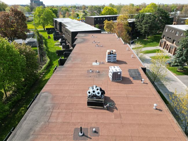 Aerial view of flat rooftops with HVAC units, surrounded by trees and nearby buildings.