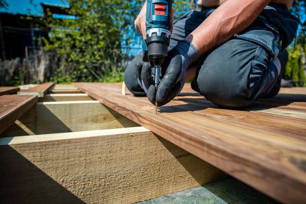 A person kneeling on a wooden deck frame, using a power drill to fasten a brown wooden plank to a support joist outdoors.
