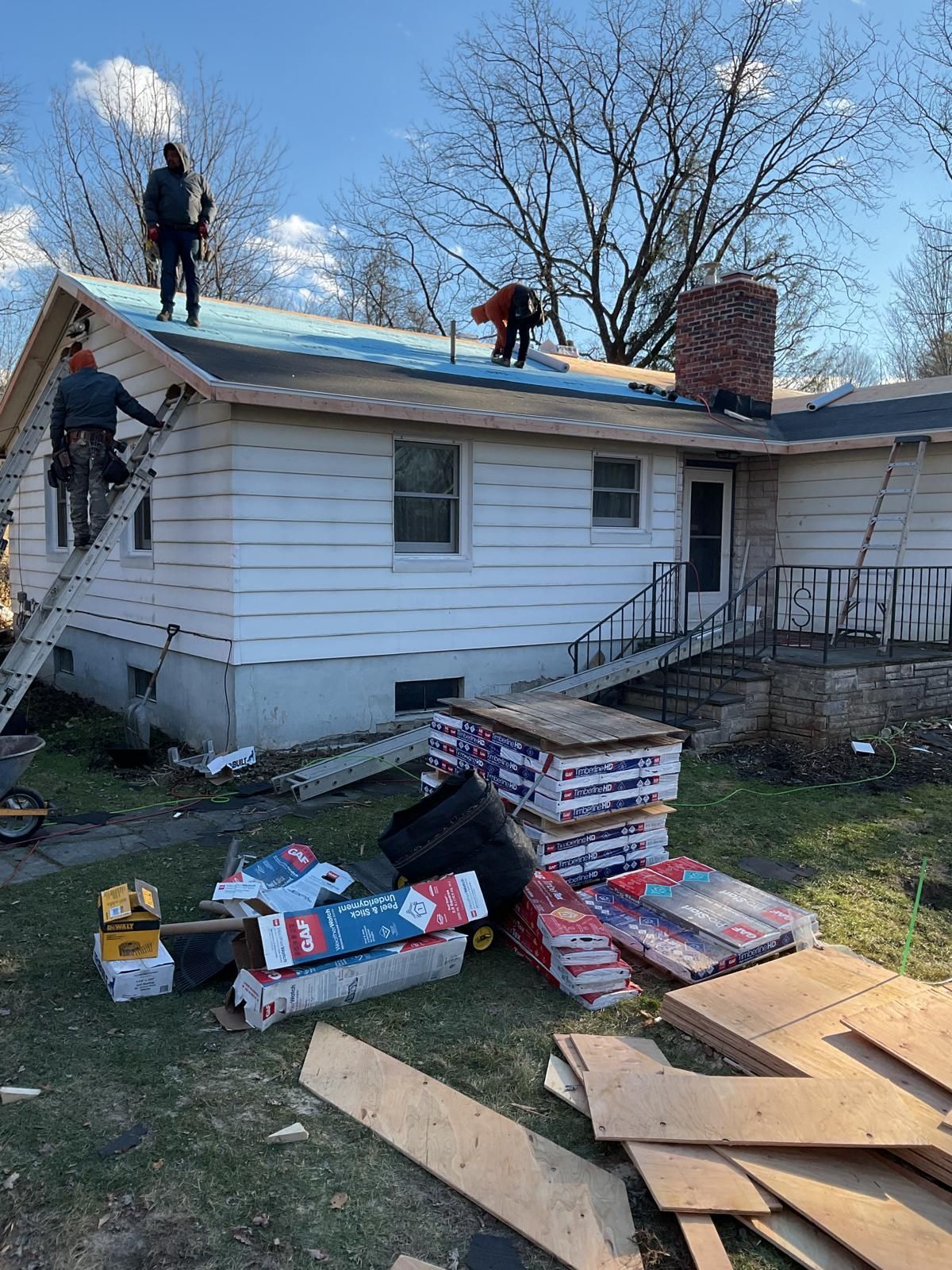 Workers replace the shingles on the roof of a white house with stacks of roofing materials in the yard below.