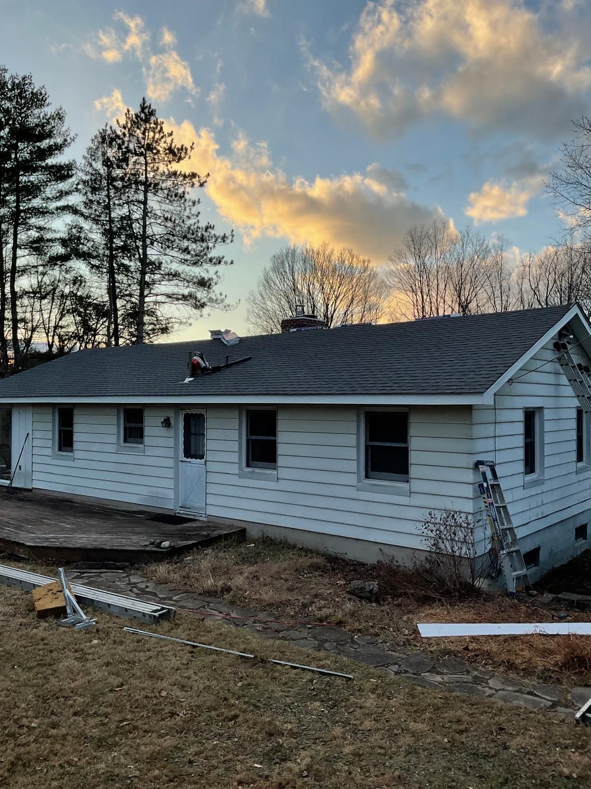 A white, single-story house with a grey shingled roof under a sunset sky, with a ladder leaning against the side.