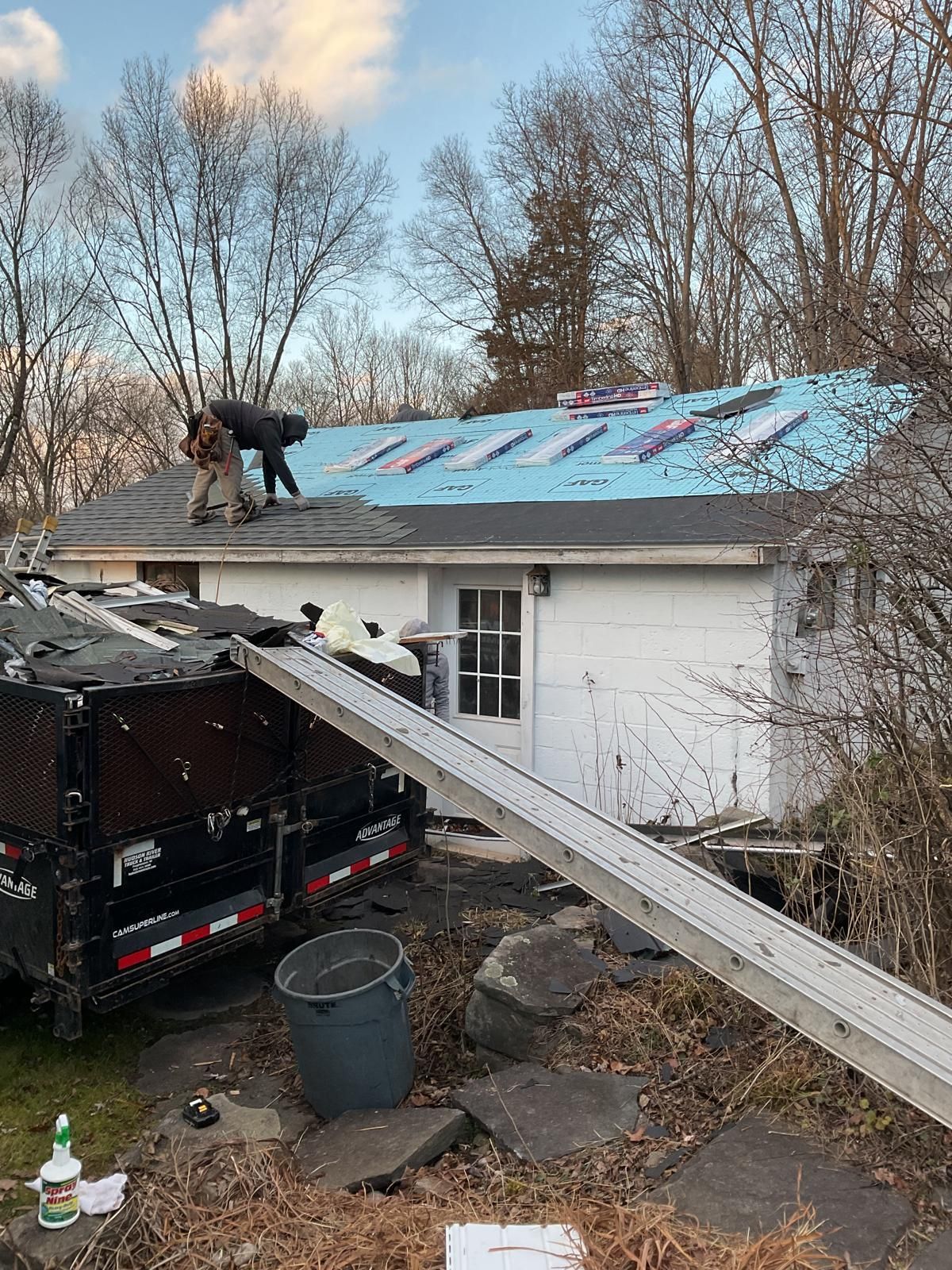 Roofers work on a house roof covered in blue underlayment, with a large debris trailer and ladder chute in the foreground.