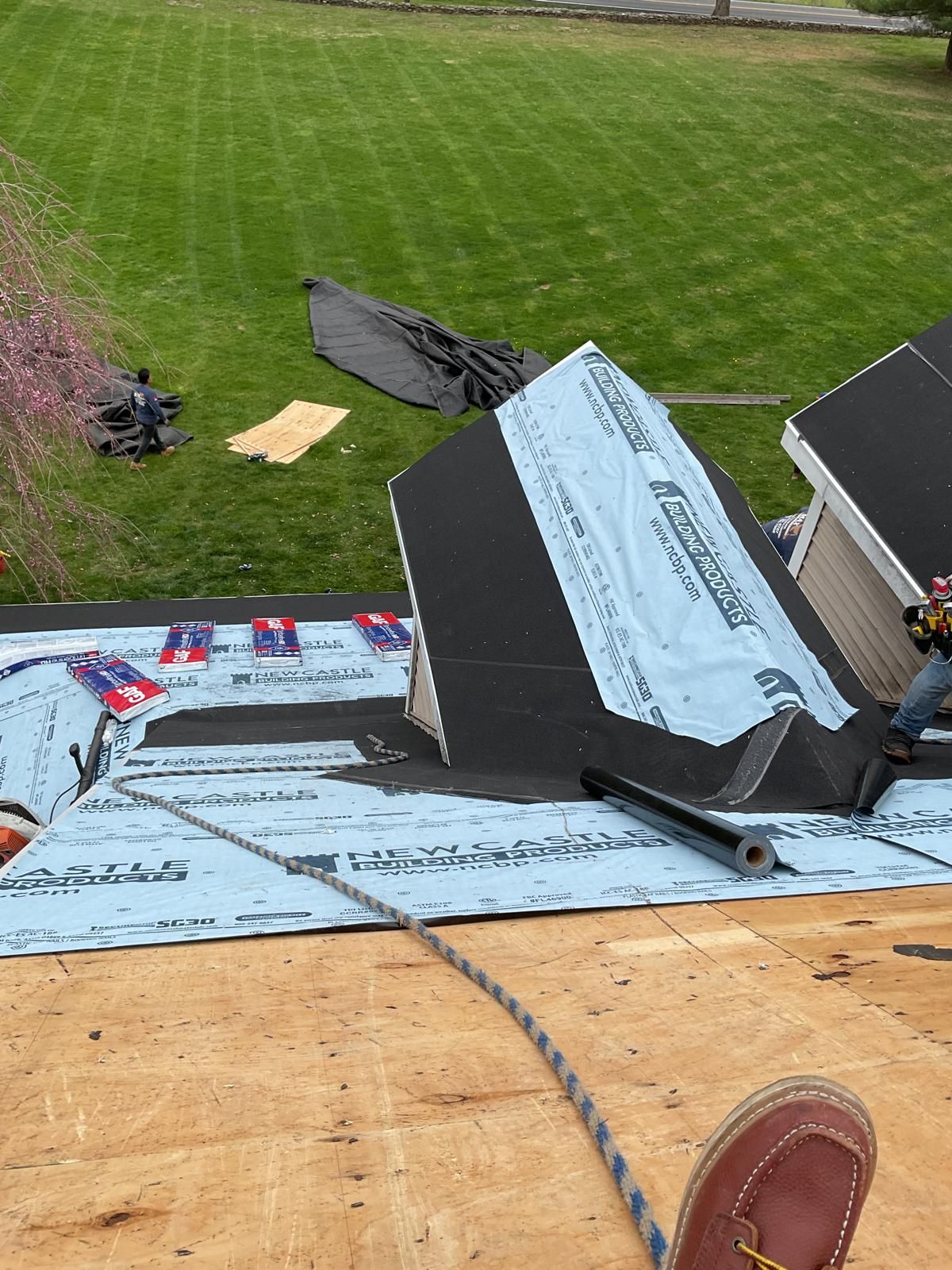 A high-angle view shows a roof under construction, featuring exposed plywood, blue underlayment, and black felt paper.