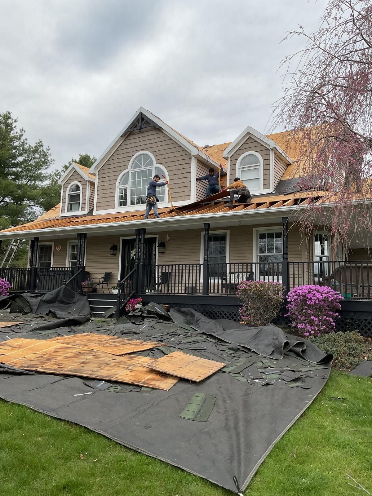 Roofers work on the shingle-stripped roof of a two-story suburban house with a wrap-around porch and debris below.