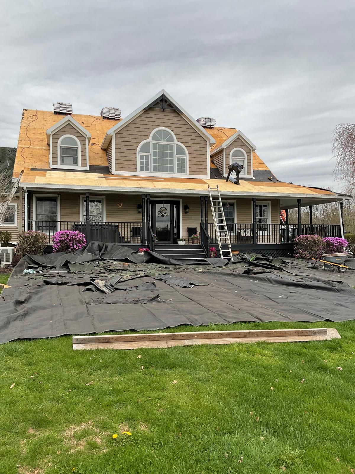Workers replace shingles on the roof of a two-story beige house with a wrap-around porch, with debris on the lawn below.