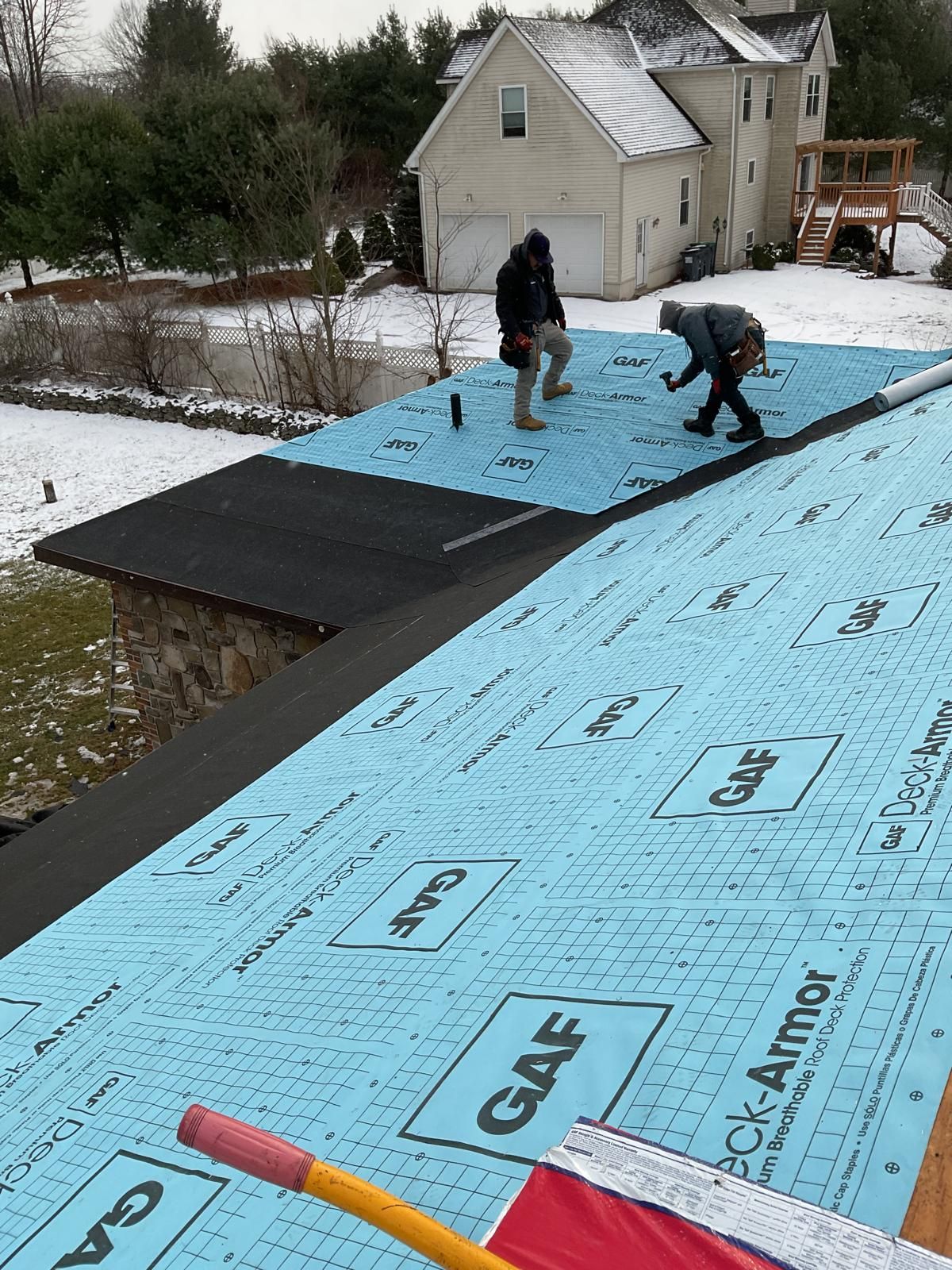 Two people in winter gear install blue GAF Deck-Armor roofing underlayment on a snowy residential roof.