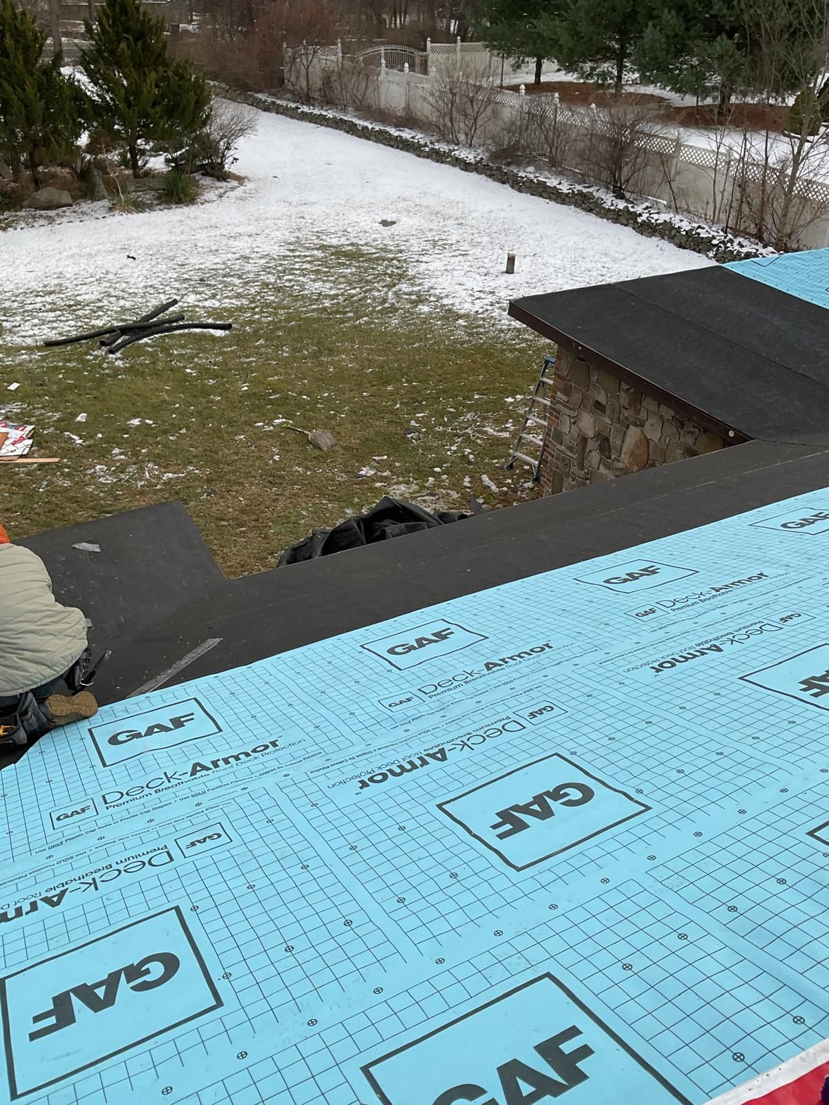 A view from a roof under construction, showing blue GAF underlayment, a worker's boots, and a snowy backyard below.