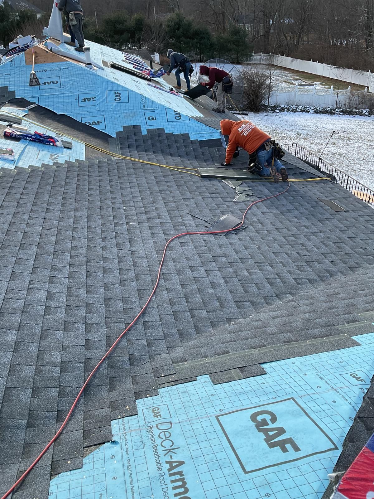 A team of roofers wearing work gear installs grey shingles on a residential roof over light blue GAF underlayment.