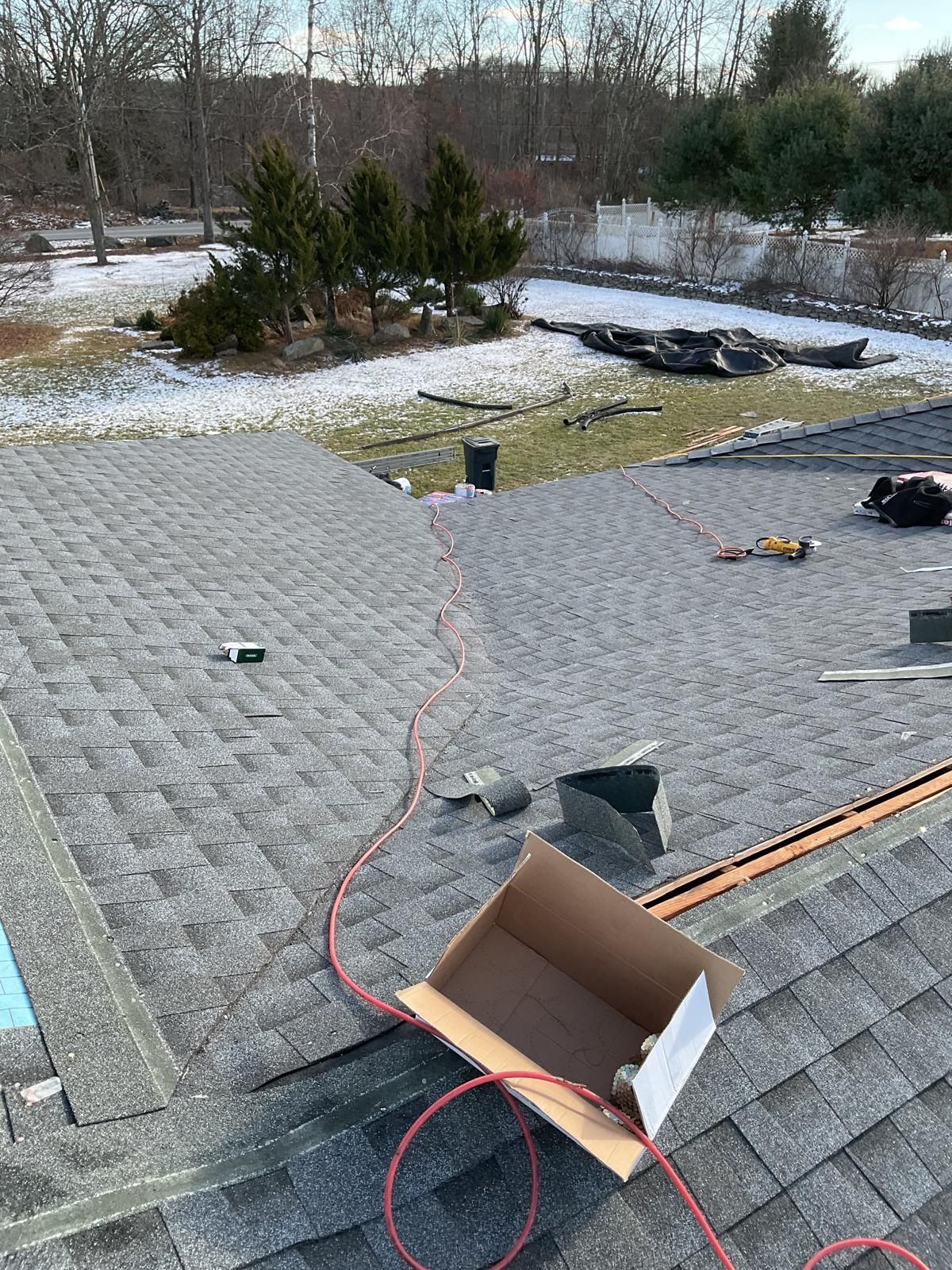 A high-angle view of a gray asphalt shingled roof under construction with an open cardboard box and red extension cords.