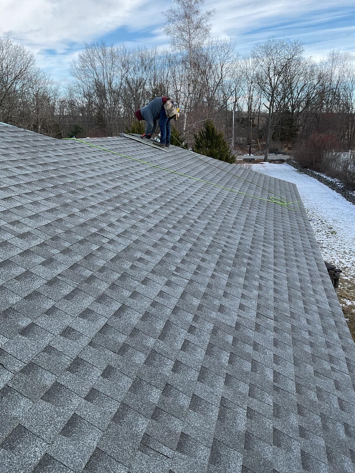 A person works on a gray shingled roof with snow on the ground in the background.