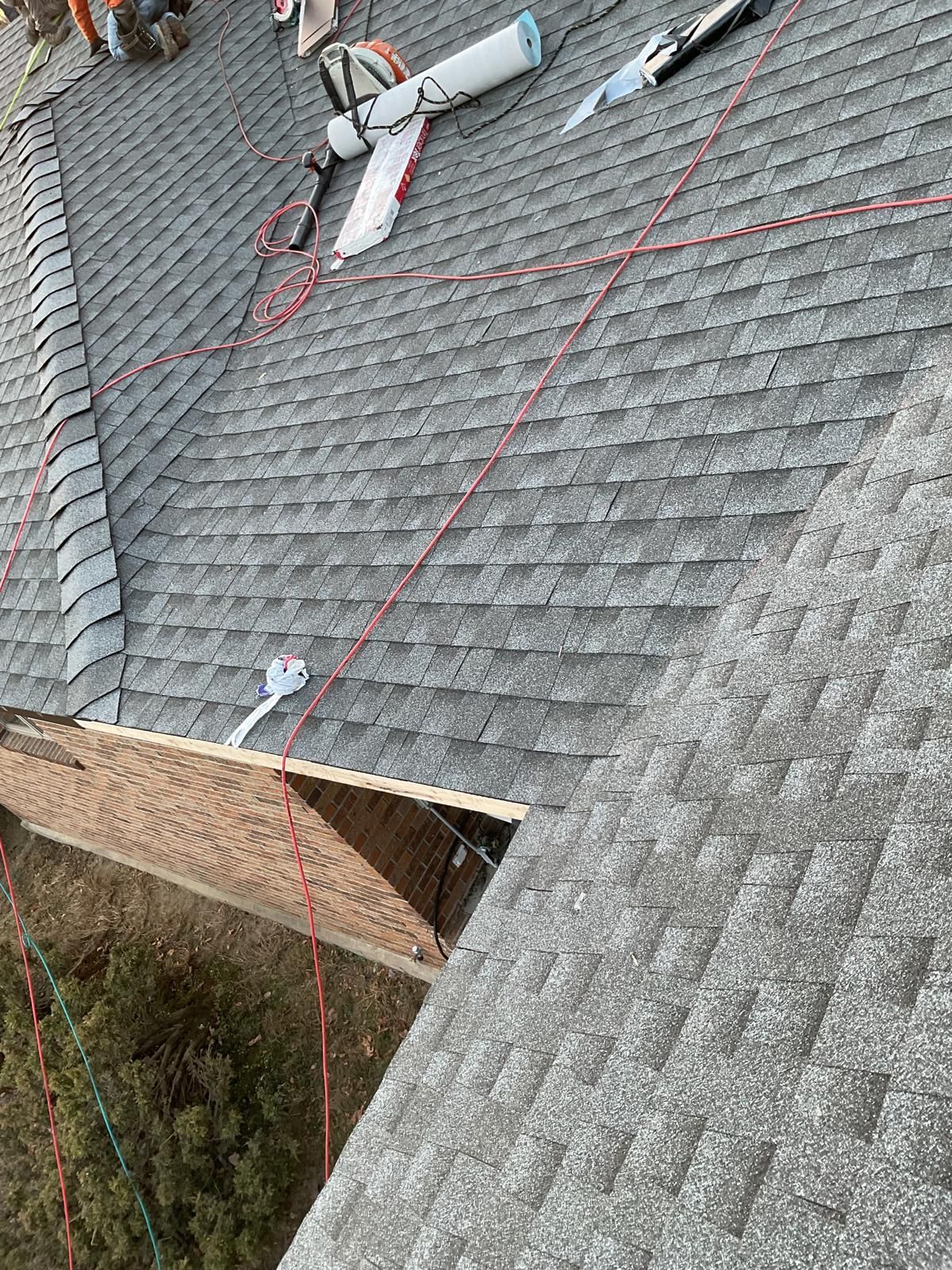 A high-angle view of a gray asphalt shingle roof under repair, with tools, red safety ropes, and exposed roof decking.