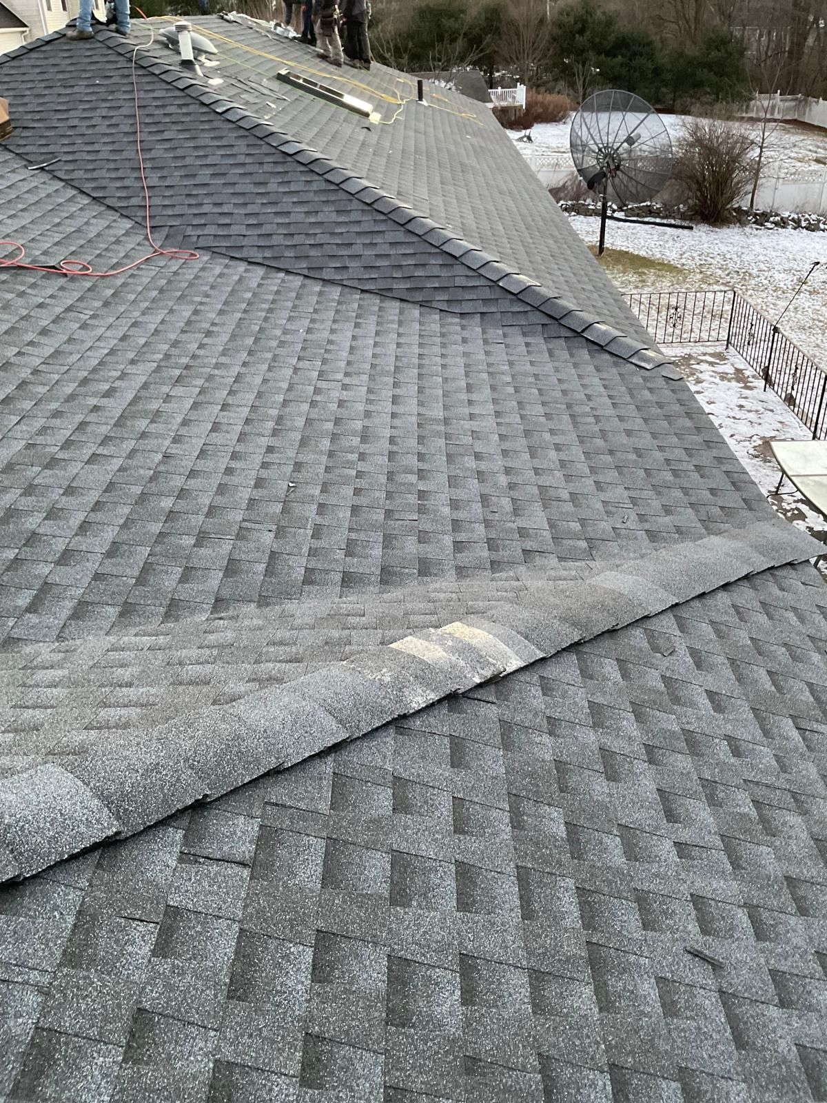High-angle view of a grey shingled roof with a diagonal ridge cap and a partially lifted shingle segment.