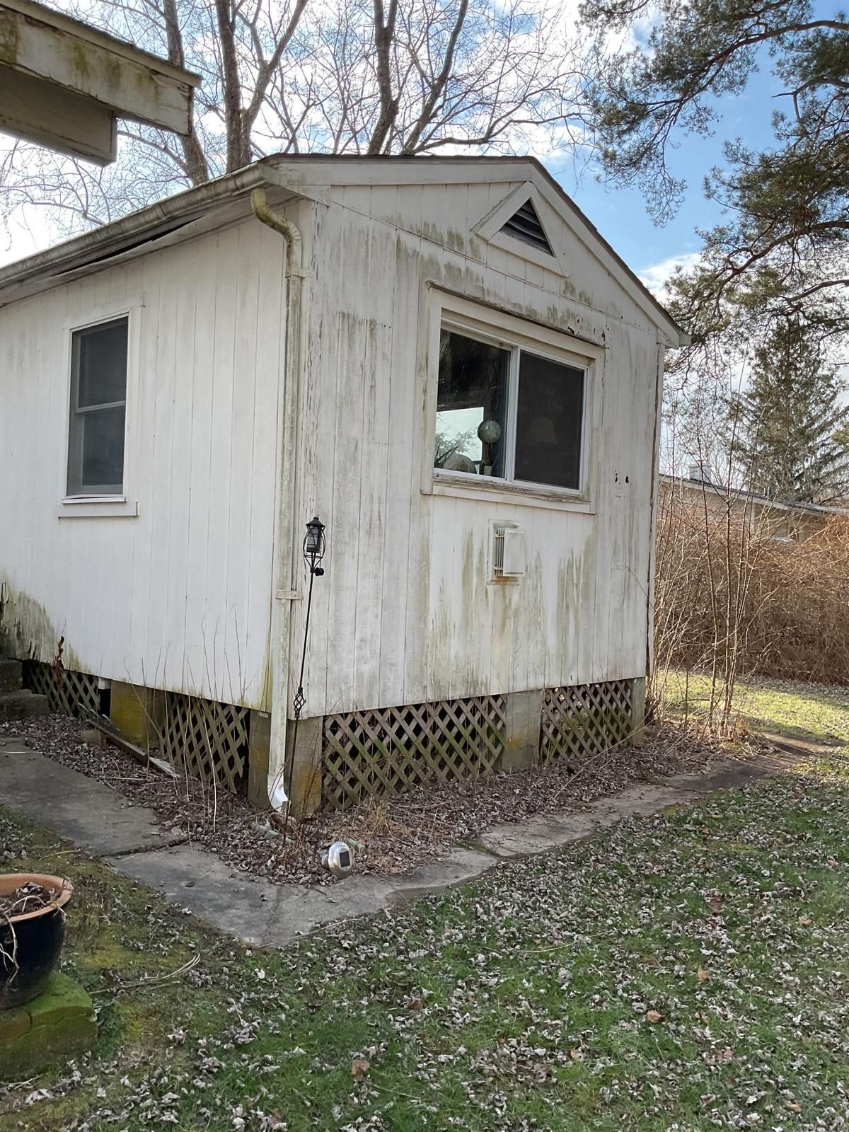 A white, weathered detached structure with a window, gabled roof, and lattice skirting, set on a lawn in late autumn.