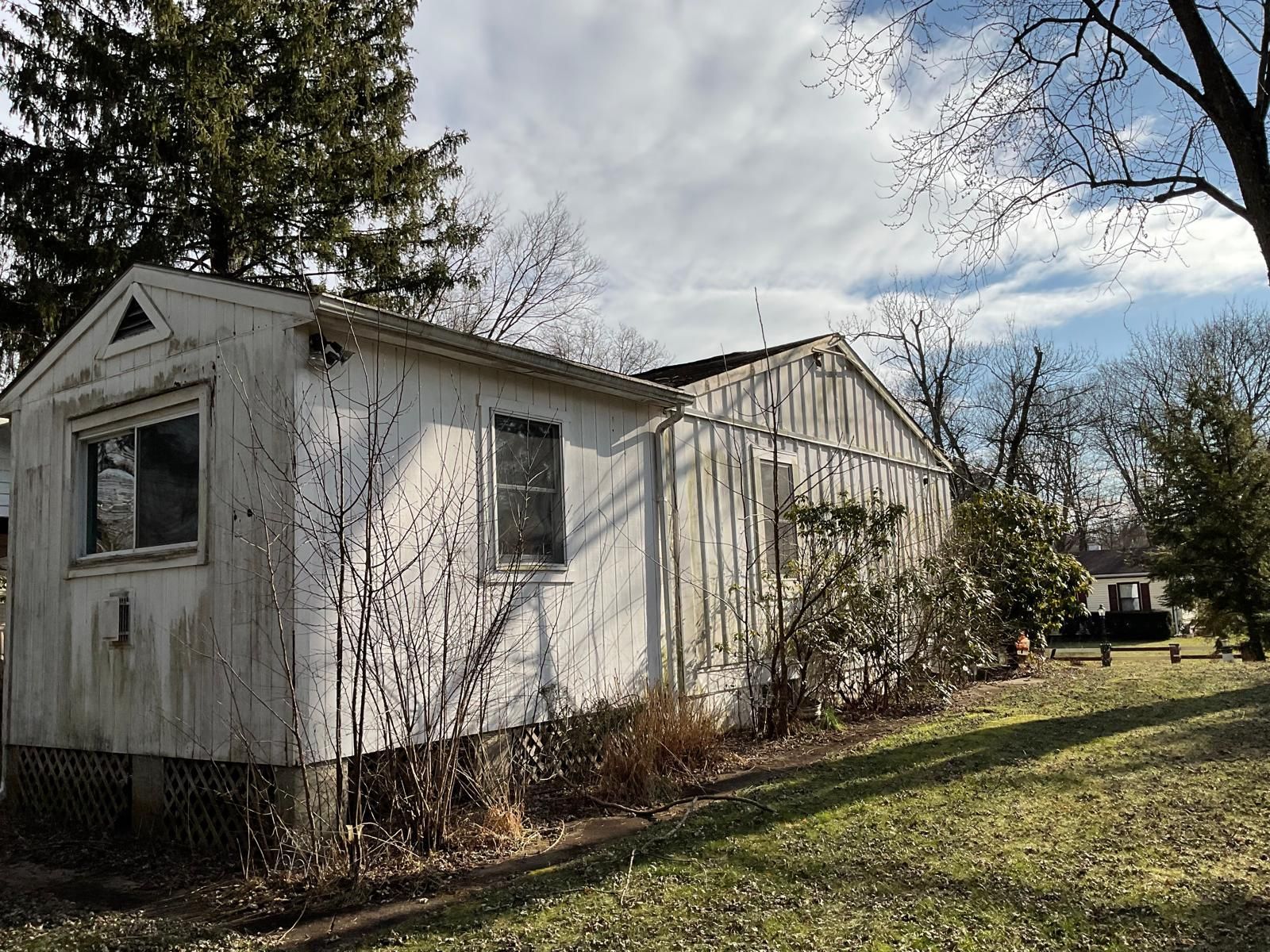 A weathered, white, single-story house with a gabled roof stands on a grassy lawn under a blue sky with scattered clouds.