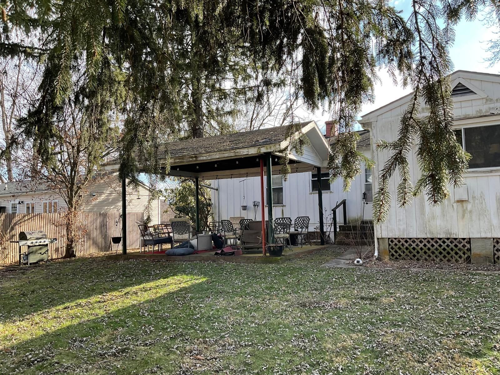 A backyard scene featuring a white house, a detached covered patio, a wooden fence, and scattered fallen leaves.