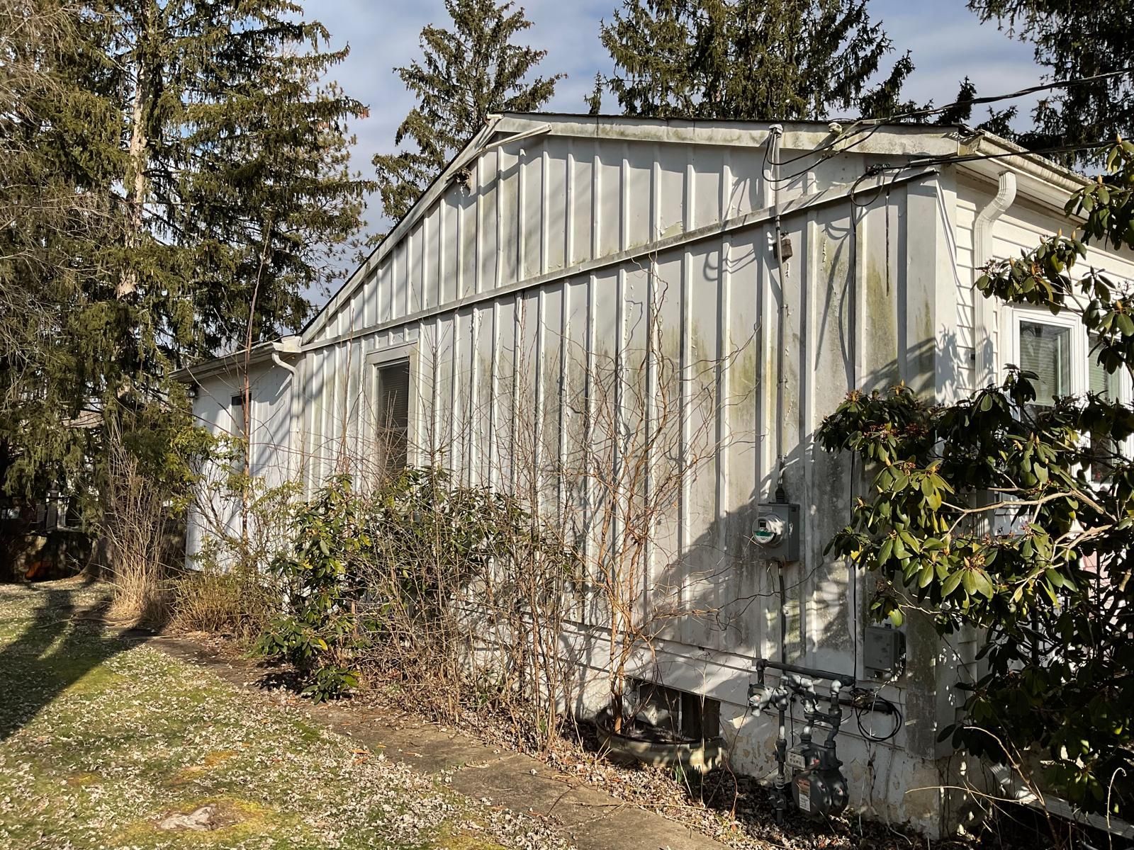 The exterior side of a weathered, white-sided house sits behind overgrown shrubs and trees on a grassy lawn.
