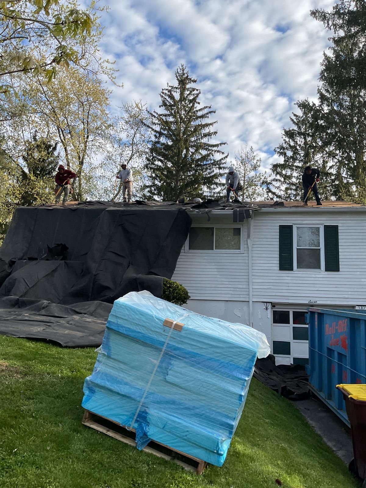 Four people are working on the roof of a white house with a blue dumpster and a pallet of blue insulation in the yard.