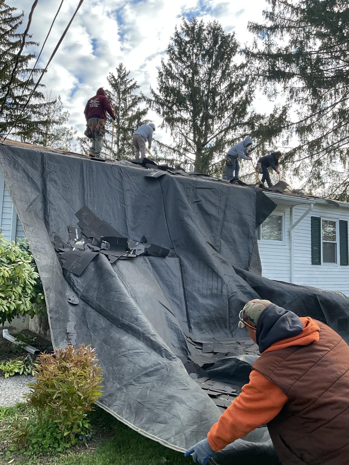 Five people working on a residential roof repair, with a large protective tarp draped over the side of the house.