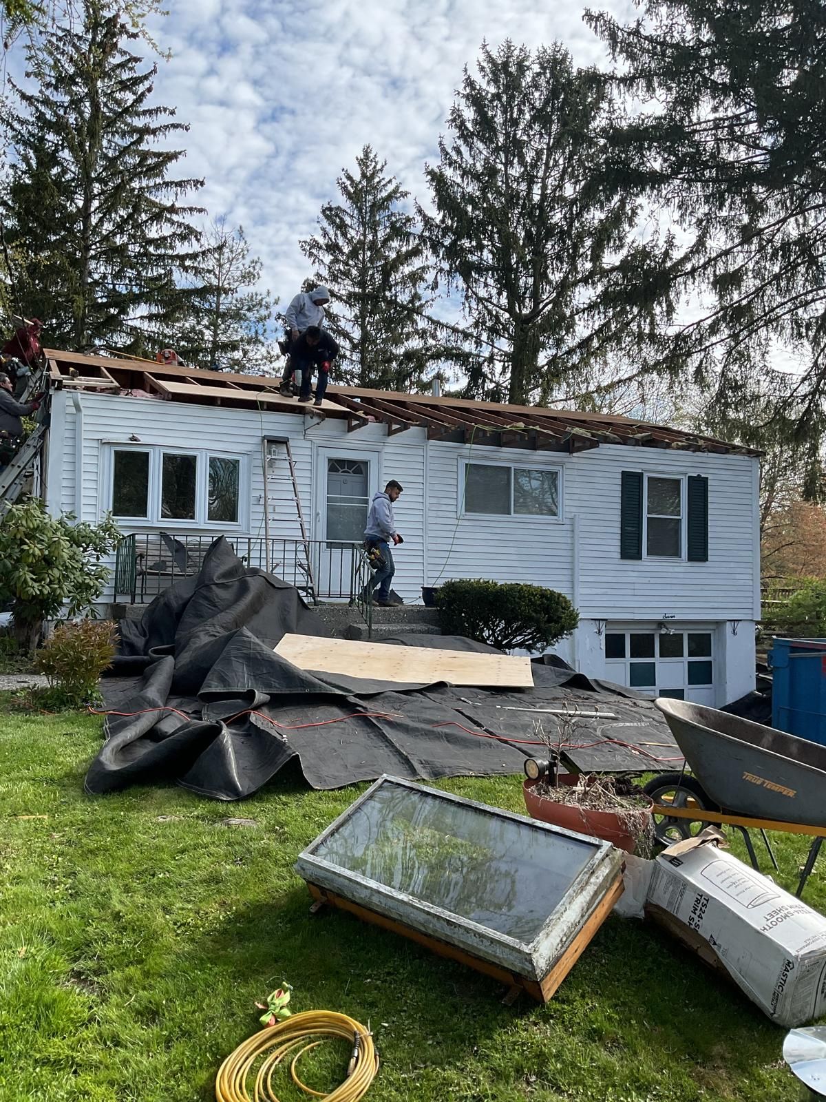 Two workers perform roof repairs on a white residential house, with debris and construction equipment on the lawn.