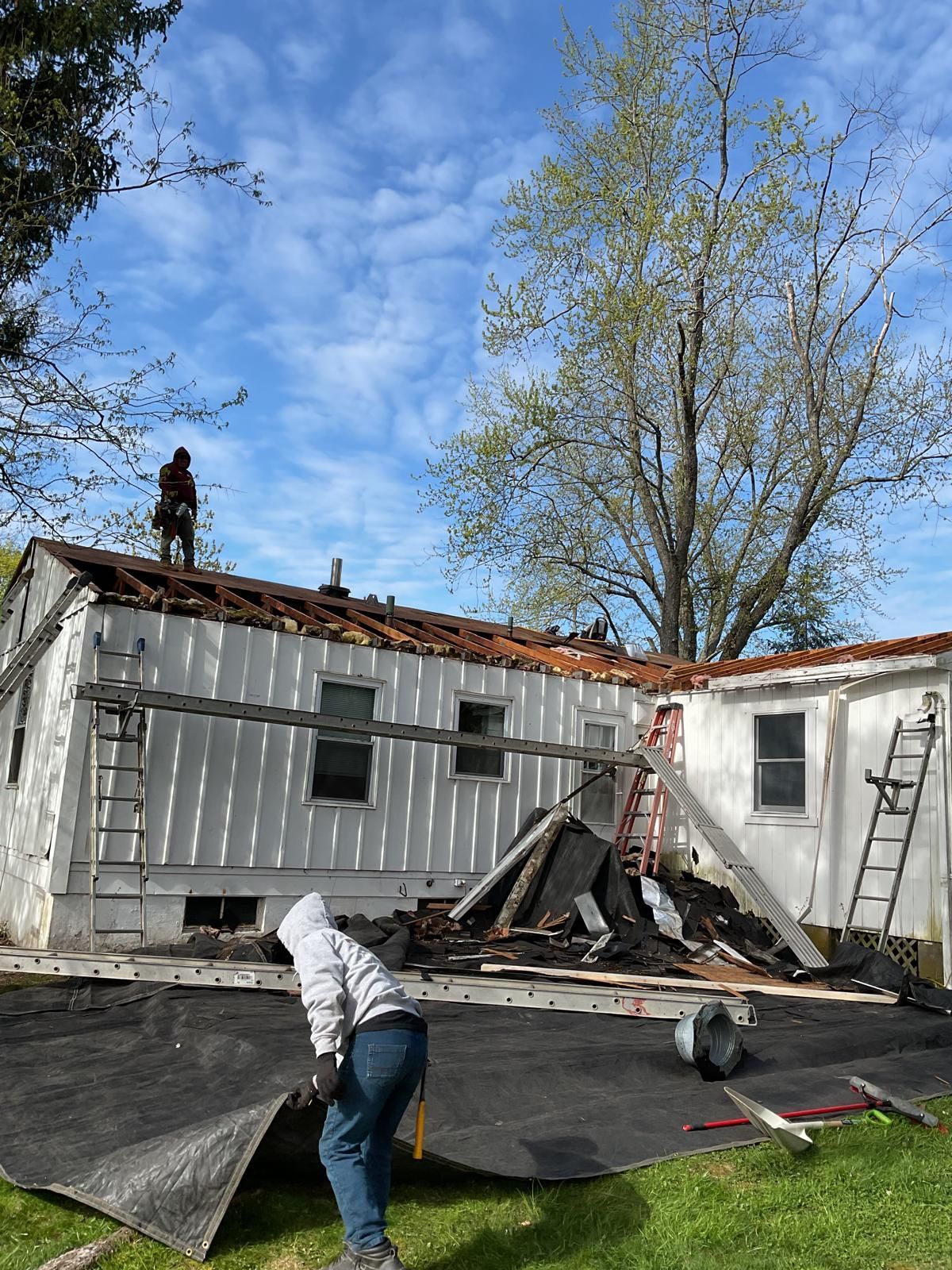 Workers remove shingles and debris from the roof of a small white house outdoors on a sunny day.