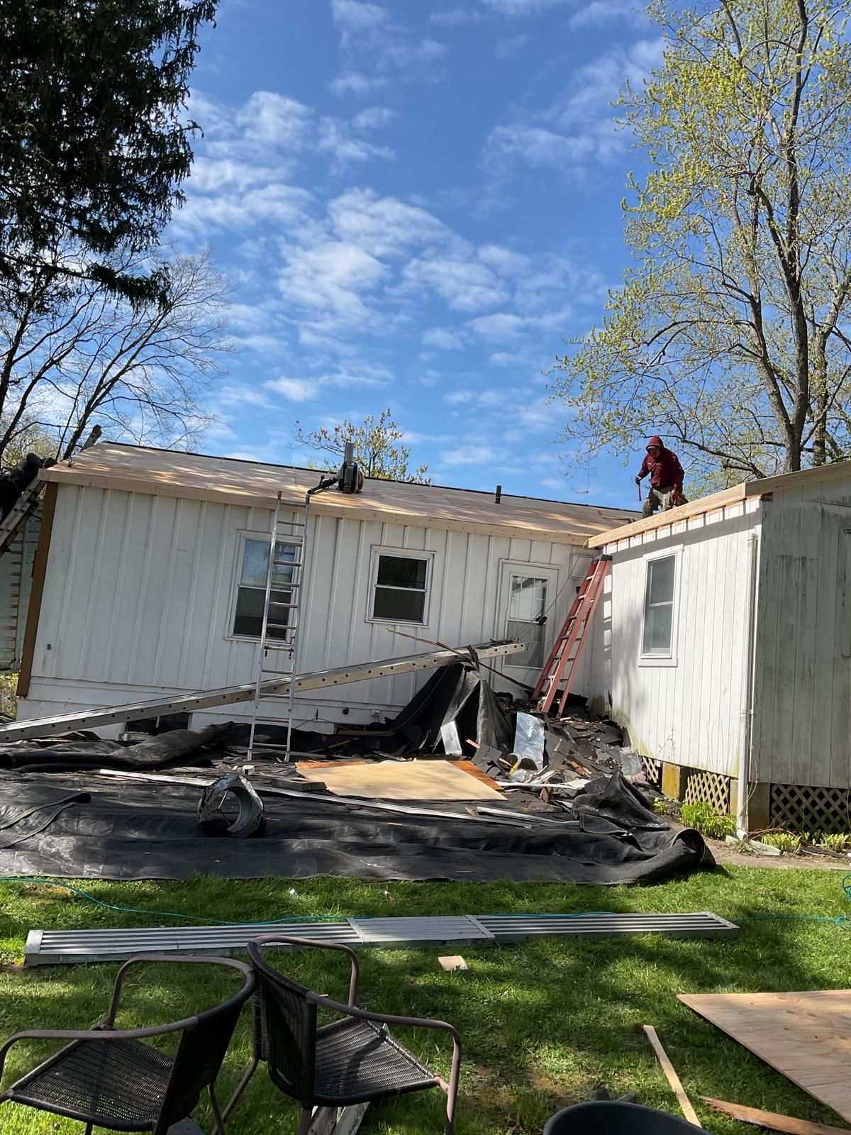 A construction worker on the roof of a white house with siding removed and debris on the lawn during a renovation.