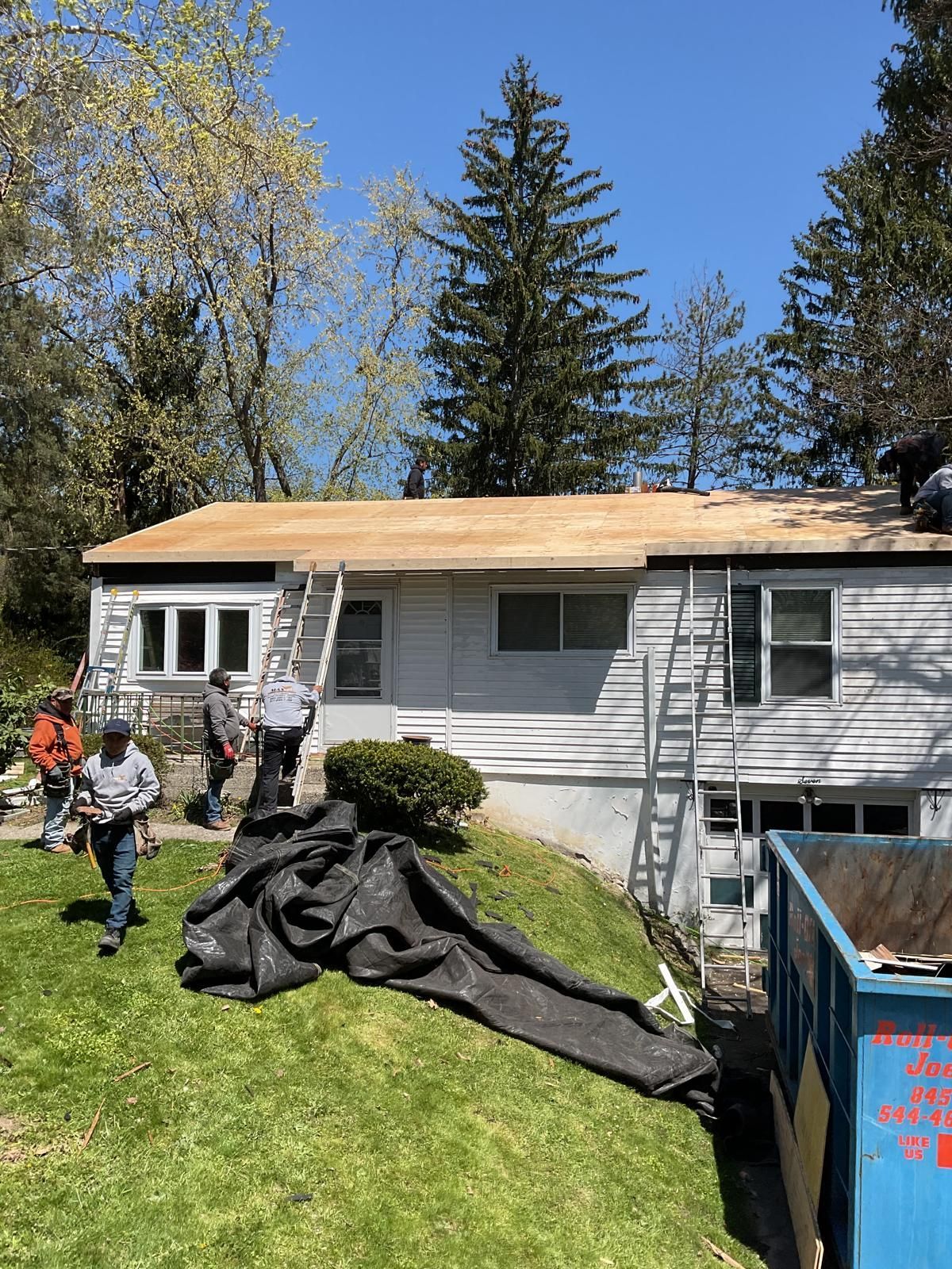 Workers repair the wooden roof of a white house with ladders and a blue dumpster nearby on a sunny day.