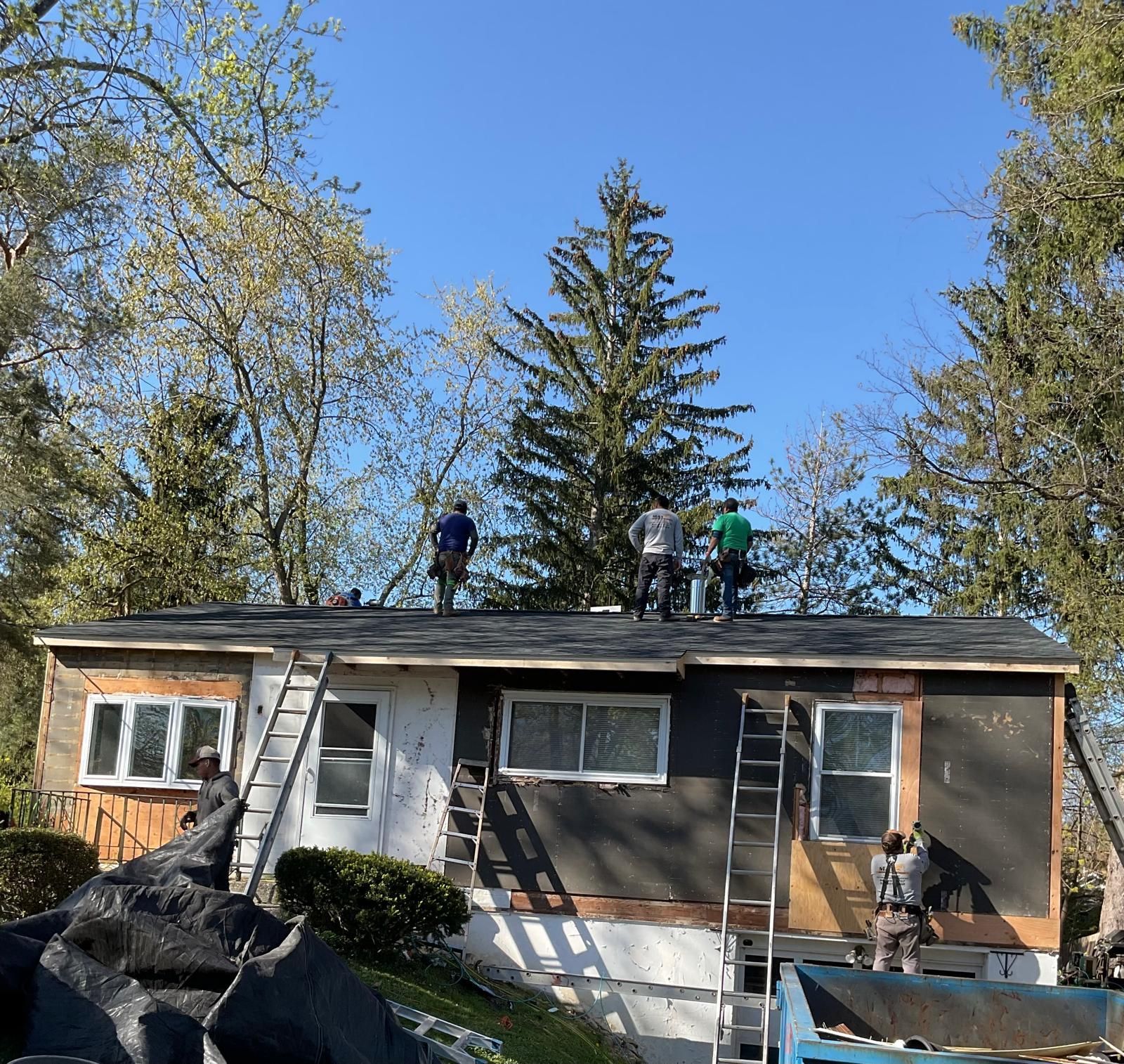 Workers in hard hats renovate the roof and exterior of a house near trees on a sunny day.