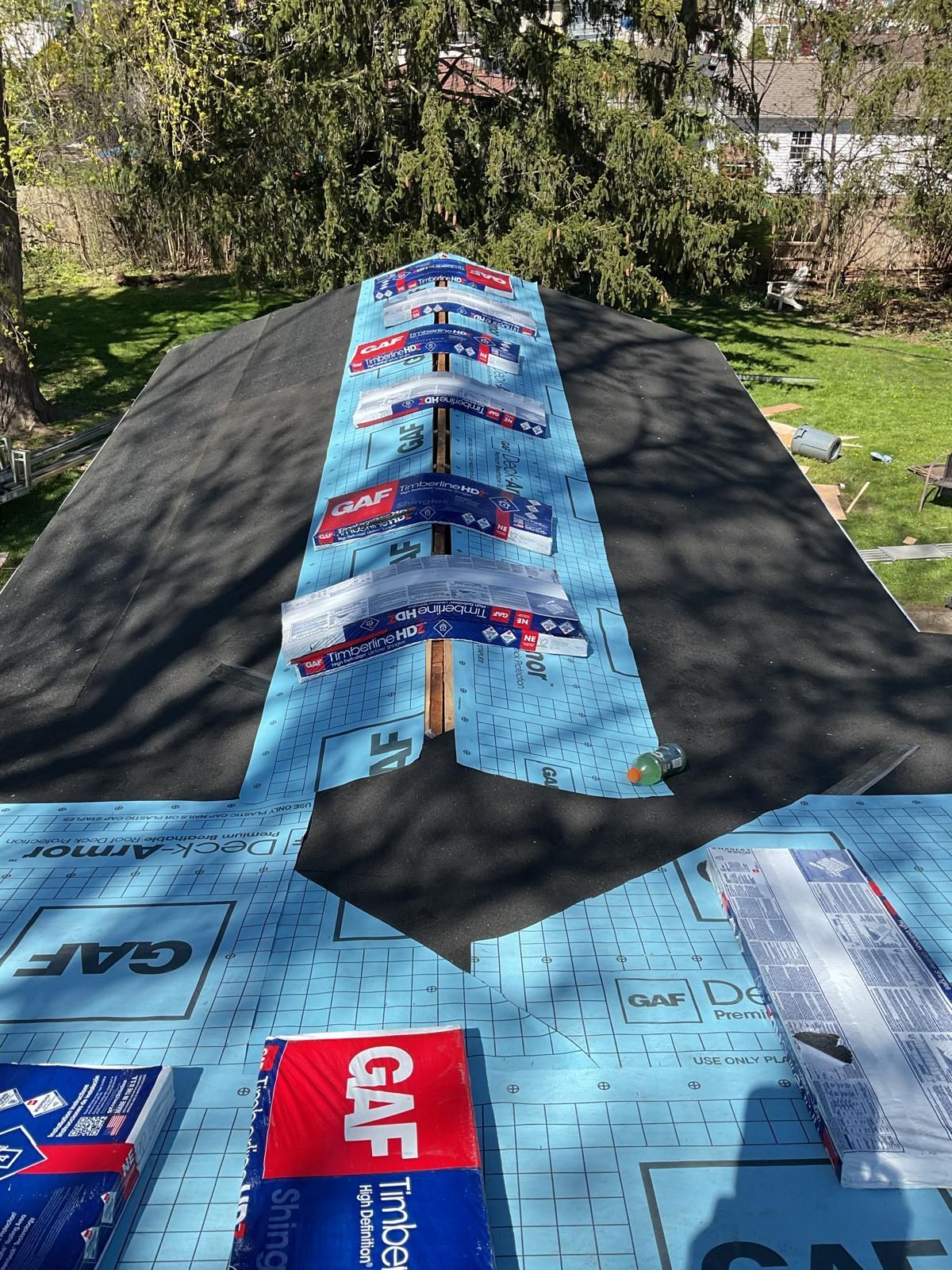 A roof under construction with blue GAF underlayment installed along the ridge and shingles stacked in the center.