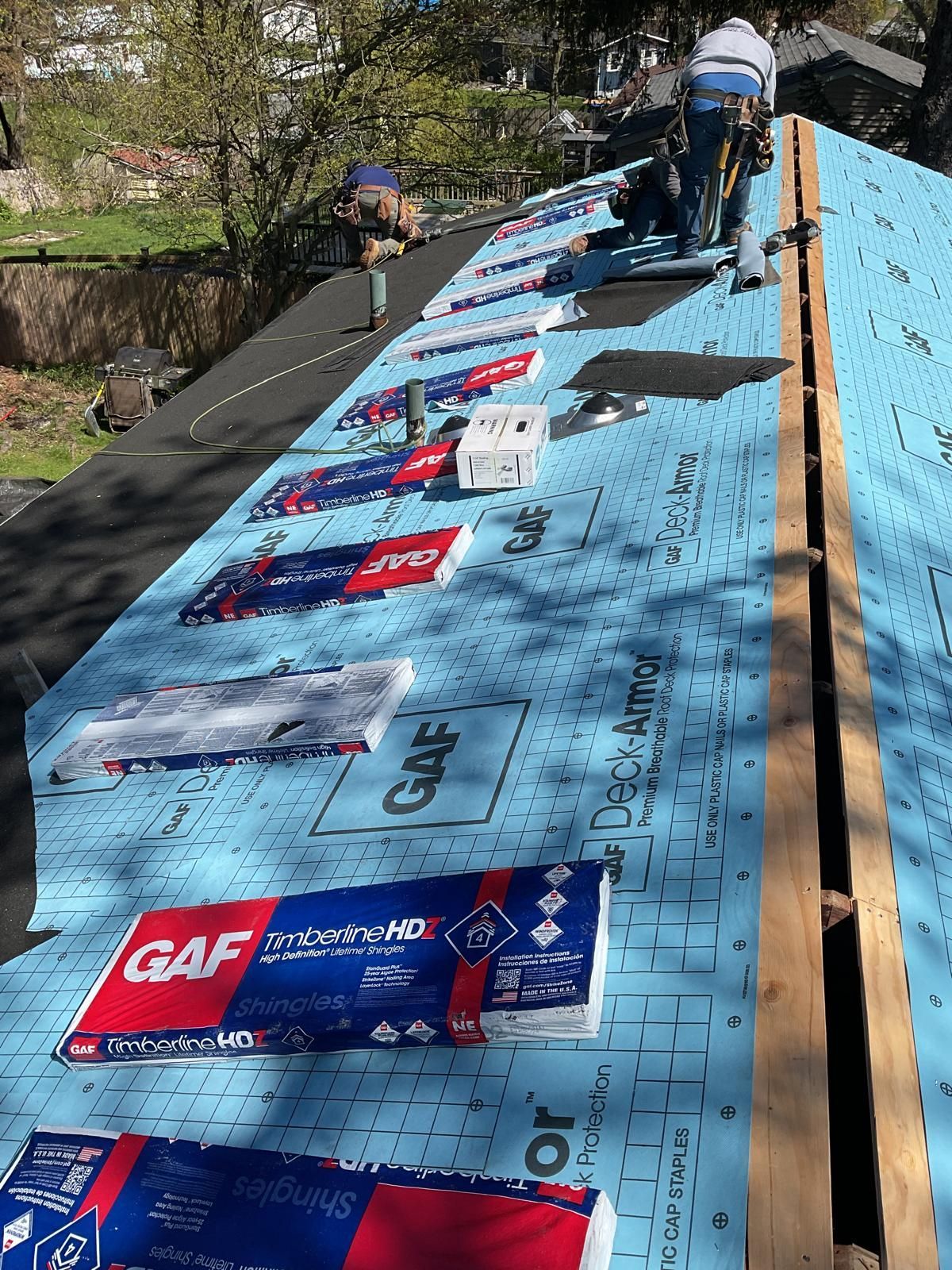 Roofers install blue GAF roofing underlayment on a steep house roof, with stacks of shingles placed along the incline.