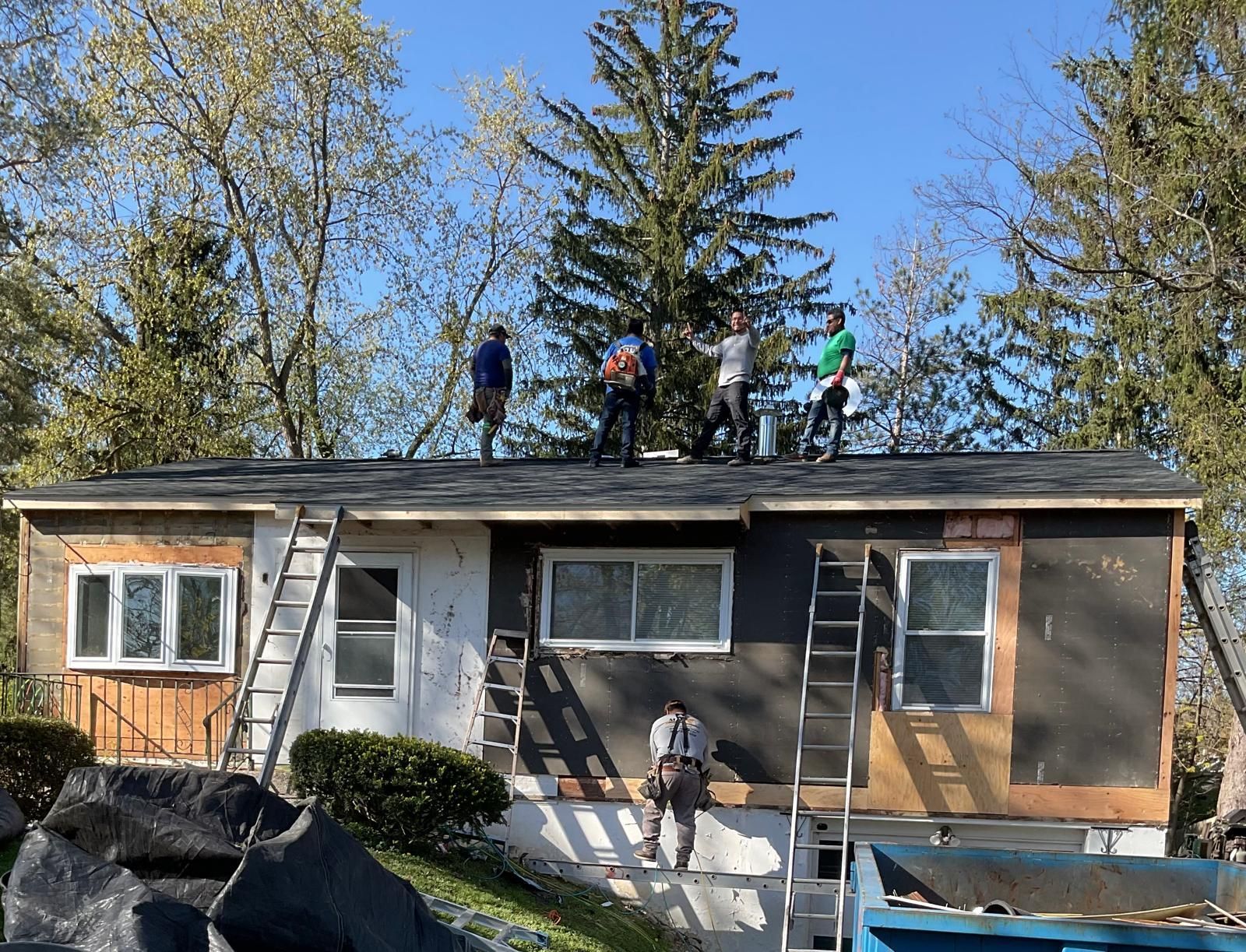 Four construction workers on the roof of a house under renovation, with ladders and a dumpster visible in the yard.