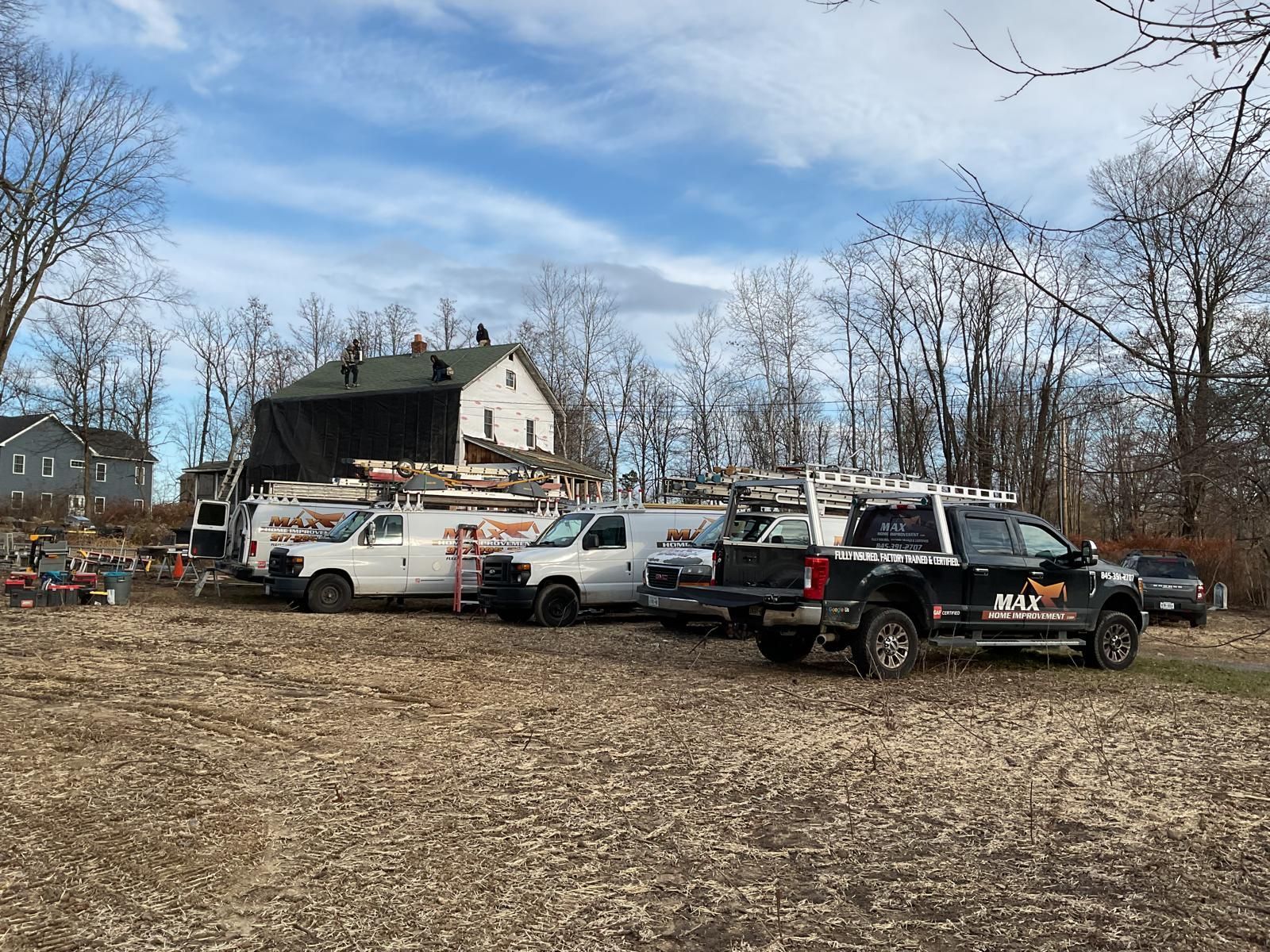 A row of service vans and trucks parked in a field near a house undergoing roof renovations under a cloudy blue sky.