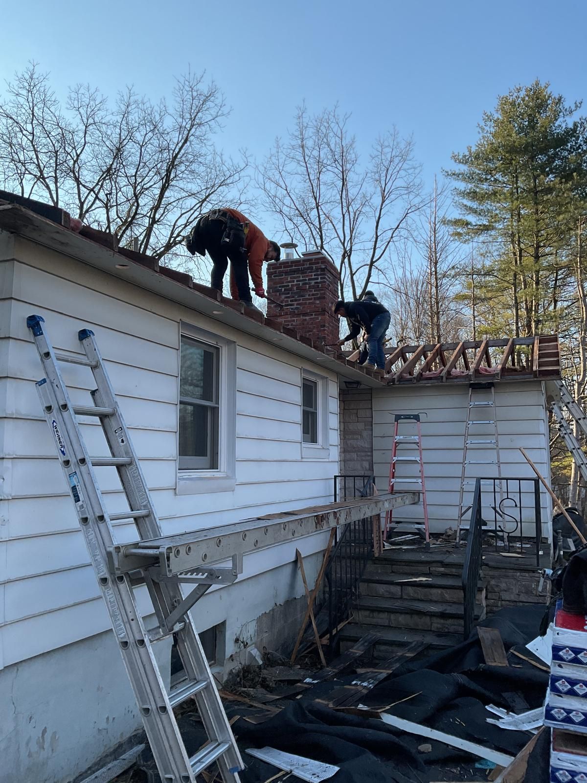 Two workers wearing high-visibility gear work on the roof and chimney of a one-story house with white siding.