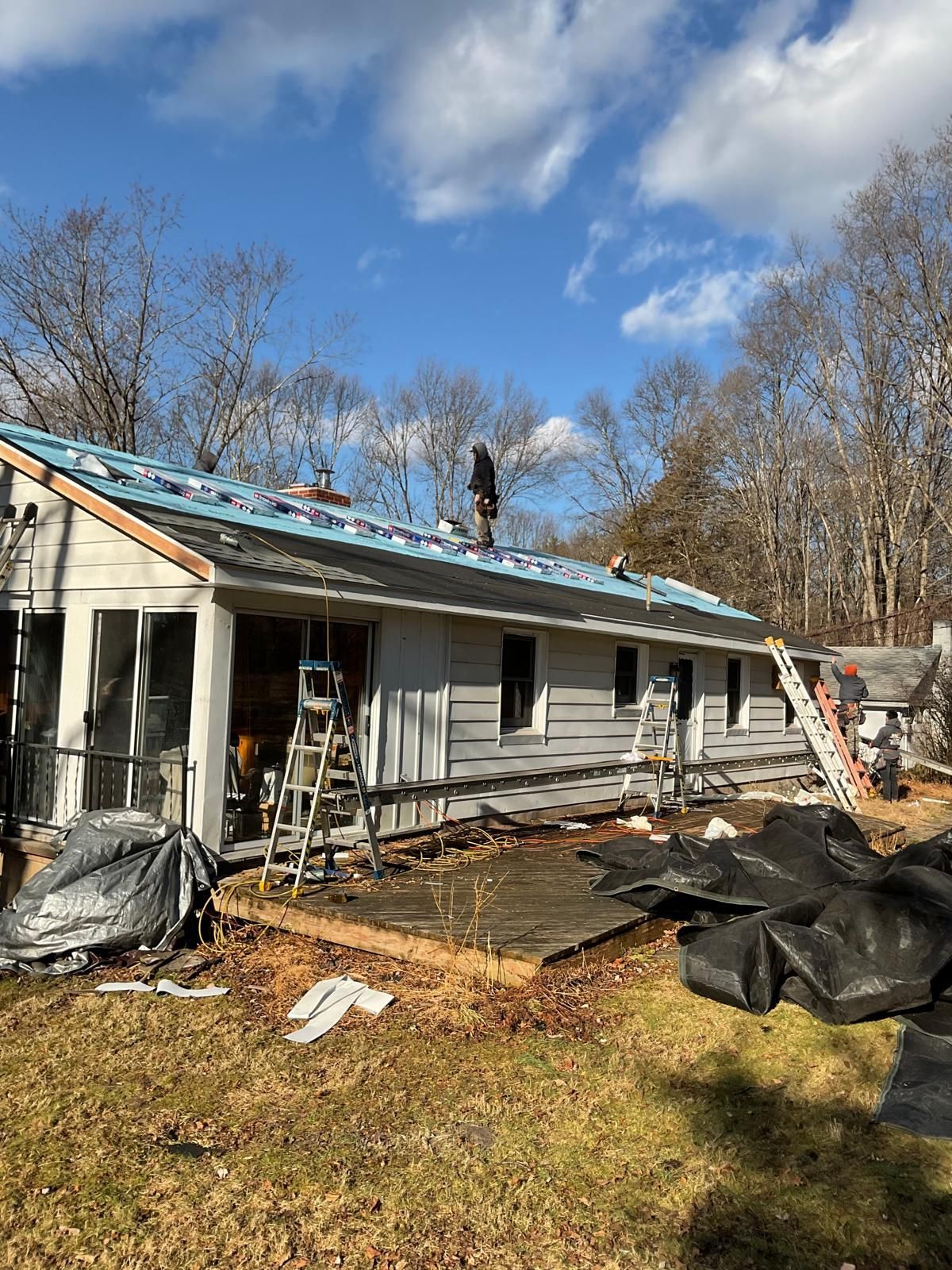 A person stands on the roof of a house under repair, with ladders leaned against the siding and discarded materials below.