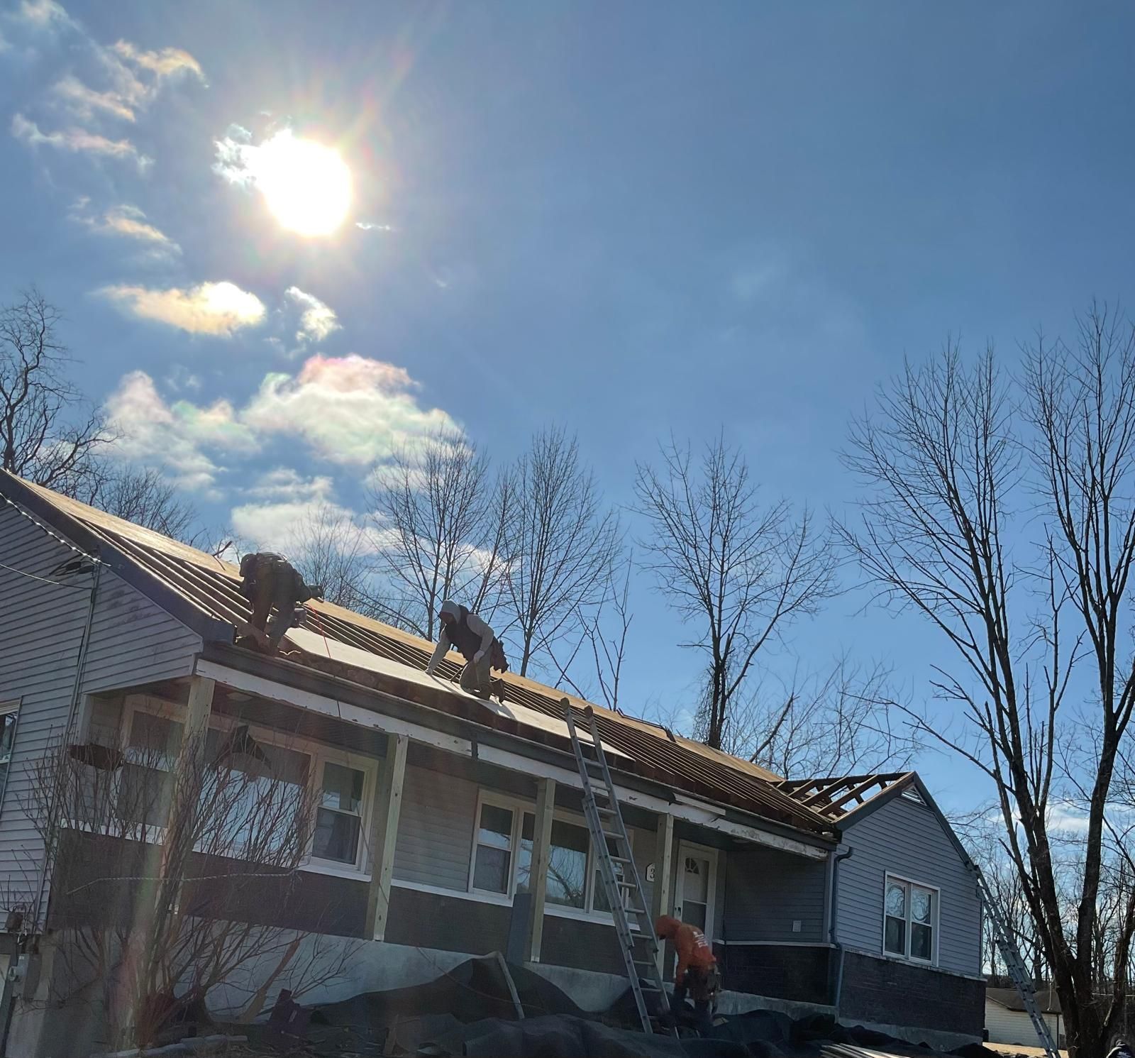 Workers on a ladder and on the roof of a house, stripping old shingles for a renovation on a sunny day.