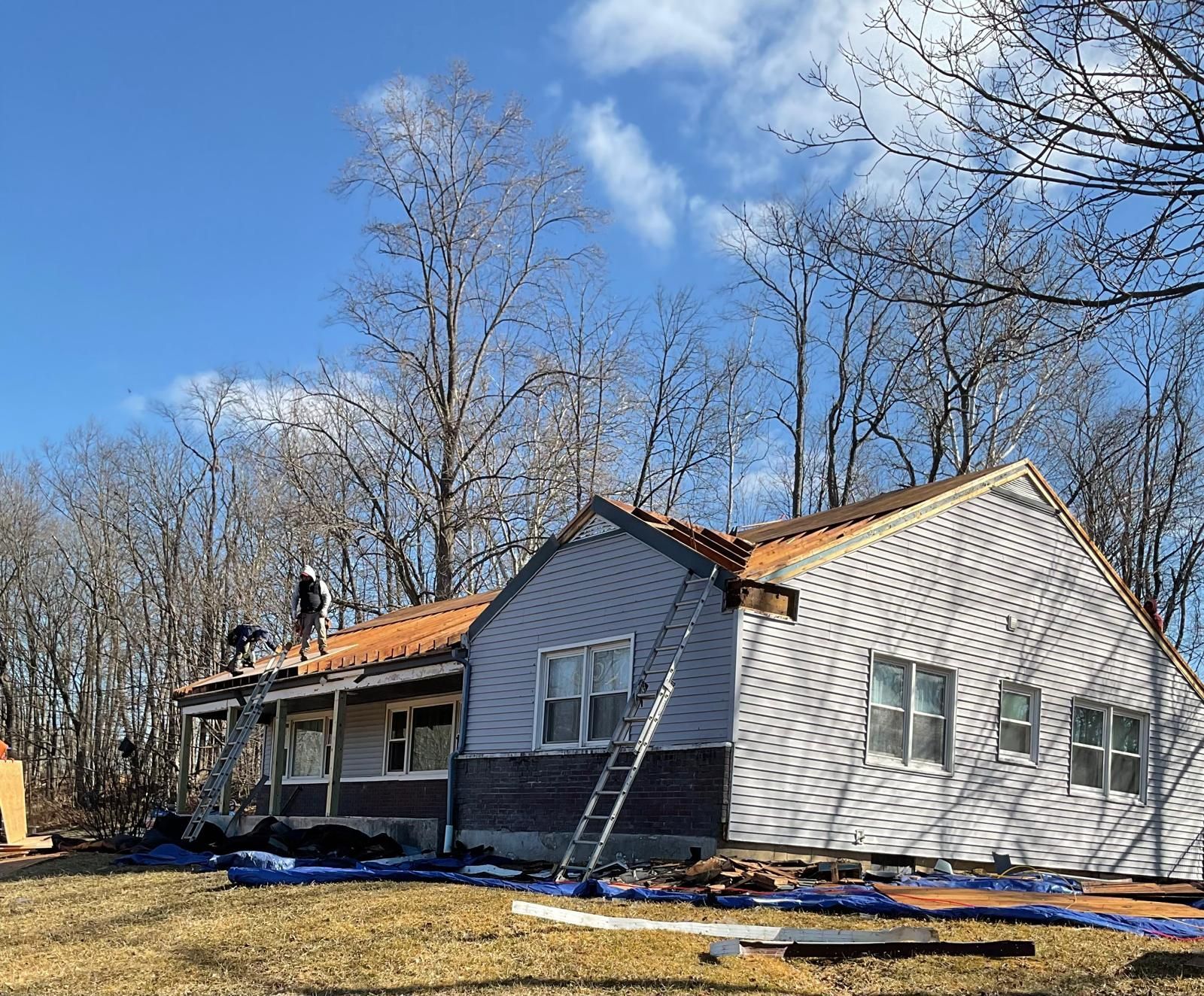 Workers replace the roof of a house on a sunny day with trees in the background.