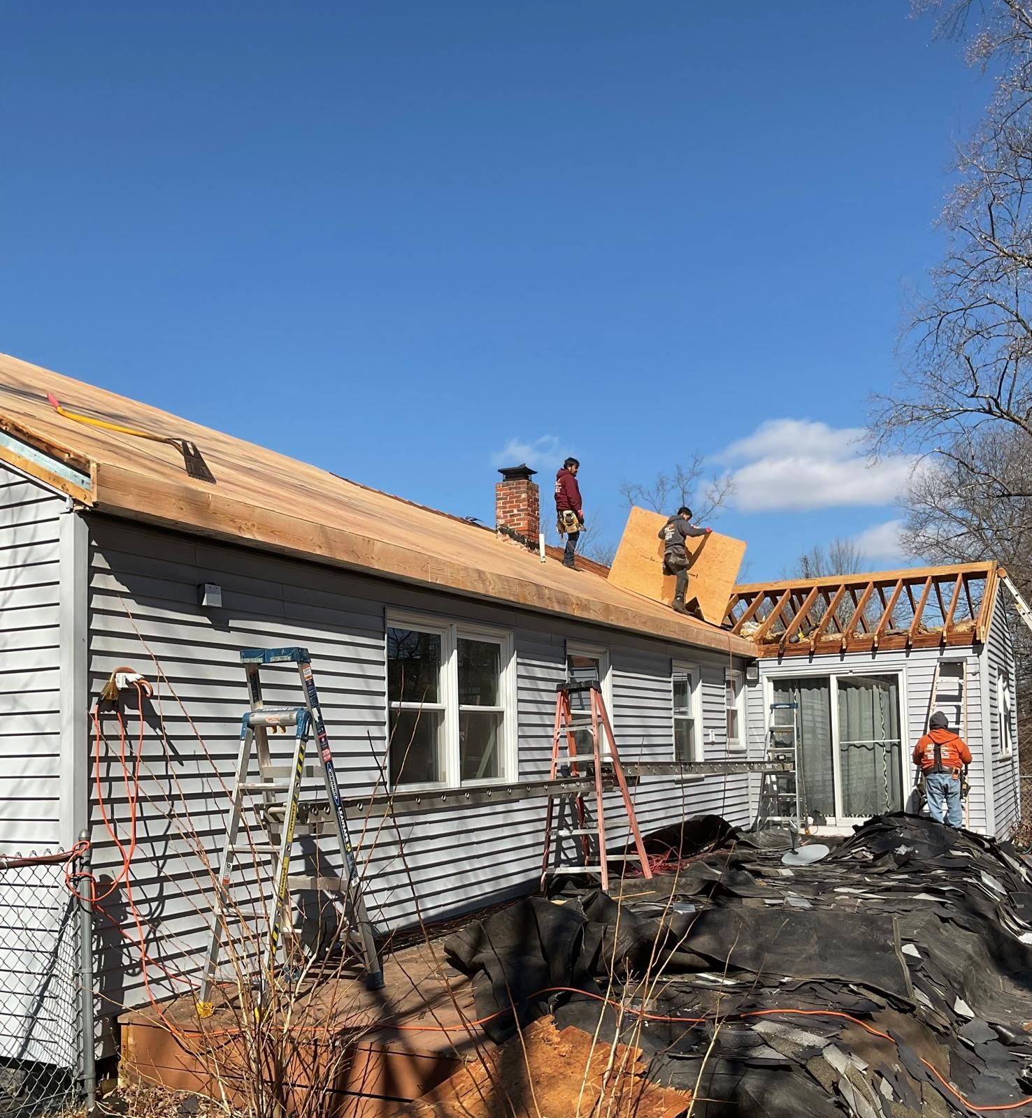 Workers installing plywood sheathing on a roof during a house renovation under a clear blue sky.