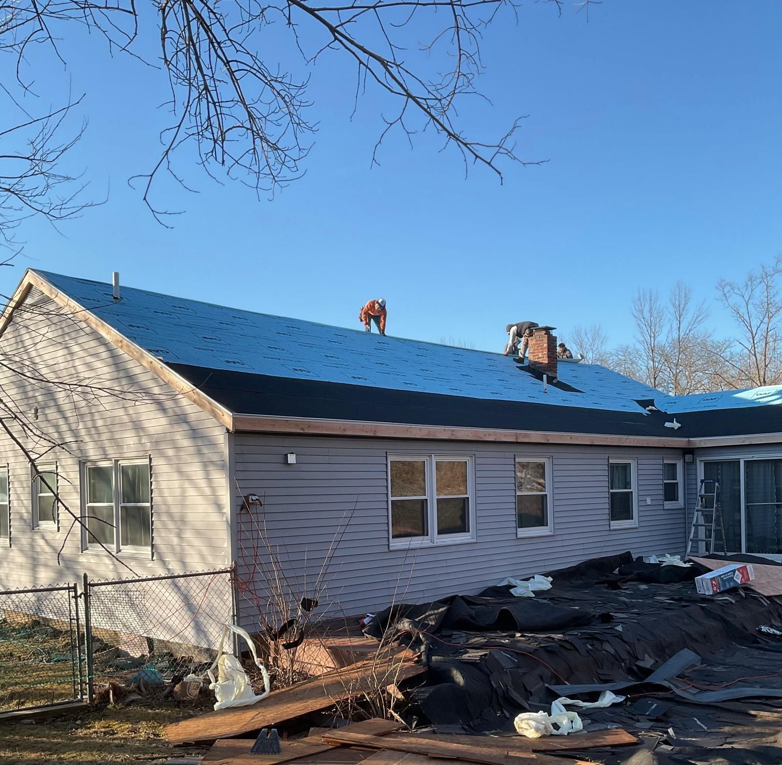 A worker in high-visibility gear installs roofing materials on a suburban house under a clear blue sky.