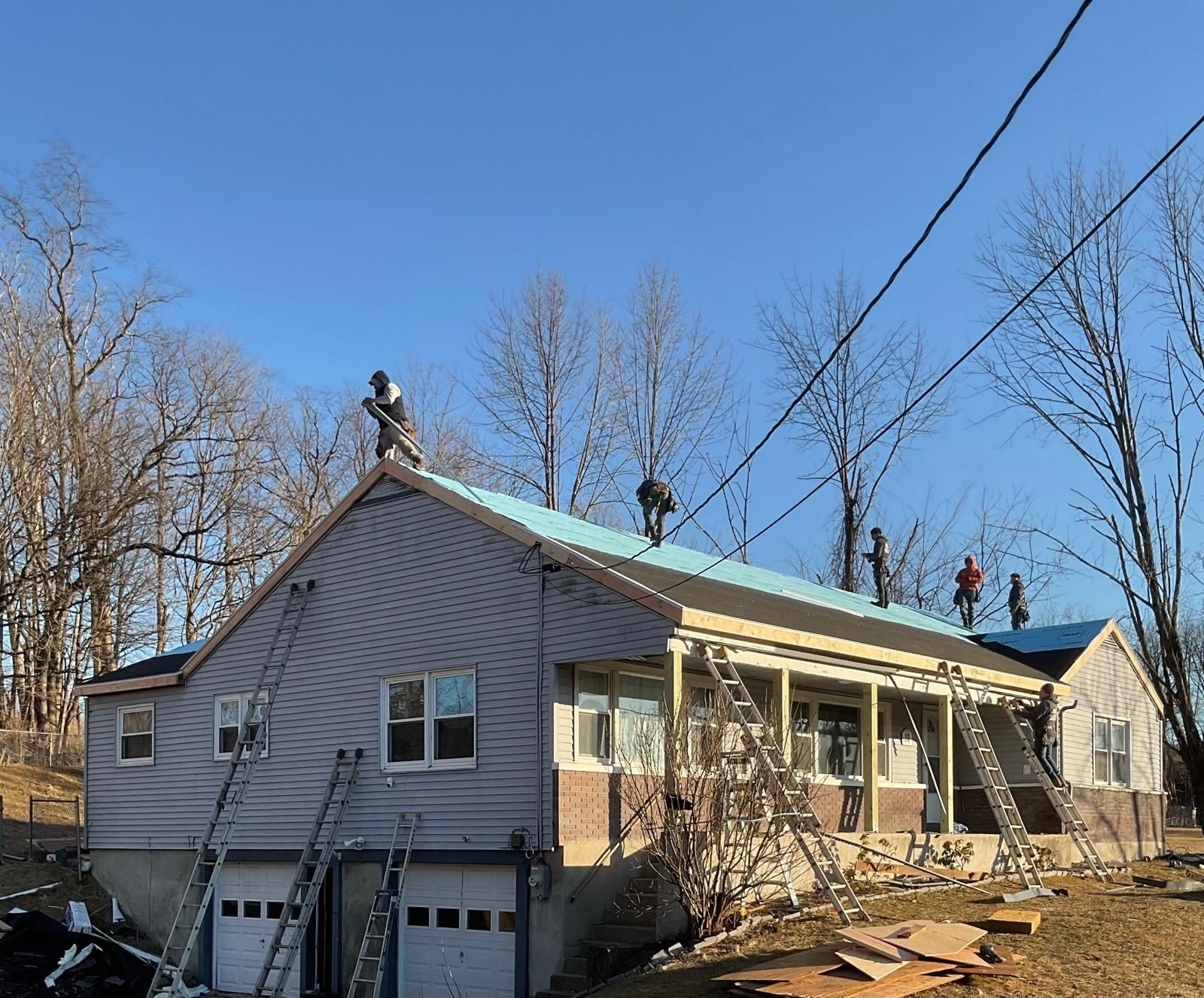 Construction workers on ladders and the roof of a house, installing new roofing material under a clear blue sky.