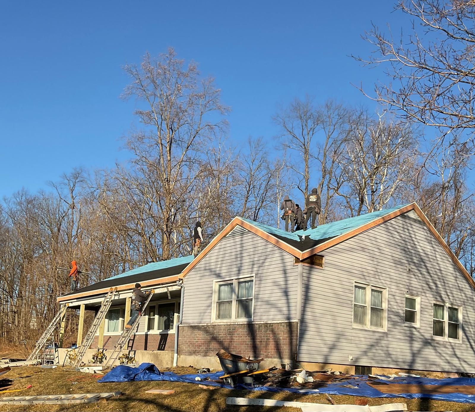 Workers on the roof of a house under construction, with a ladder leaning against the side and trees in the background.