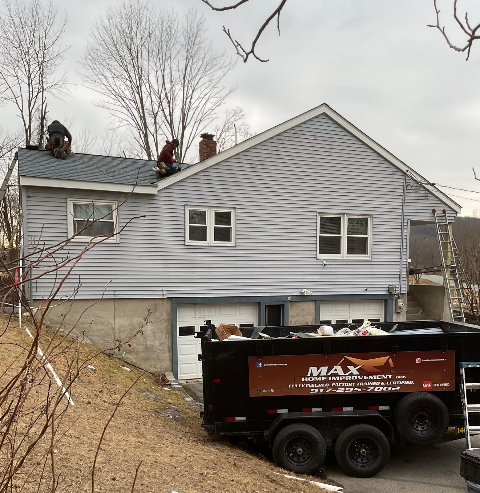 Workers replace shingles on the roof of a house with light gray siding, positioned above a parked Max dump trailer.