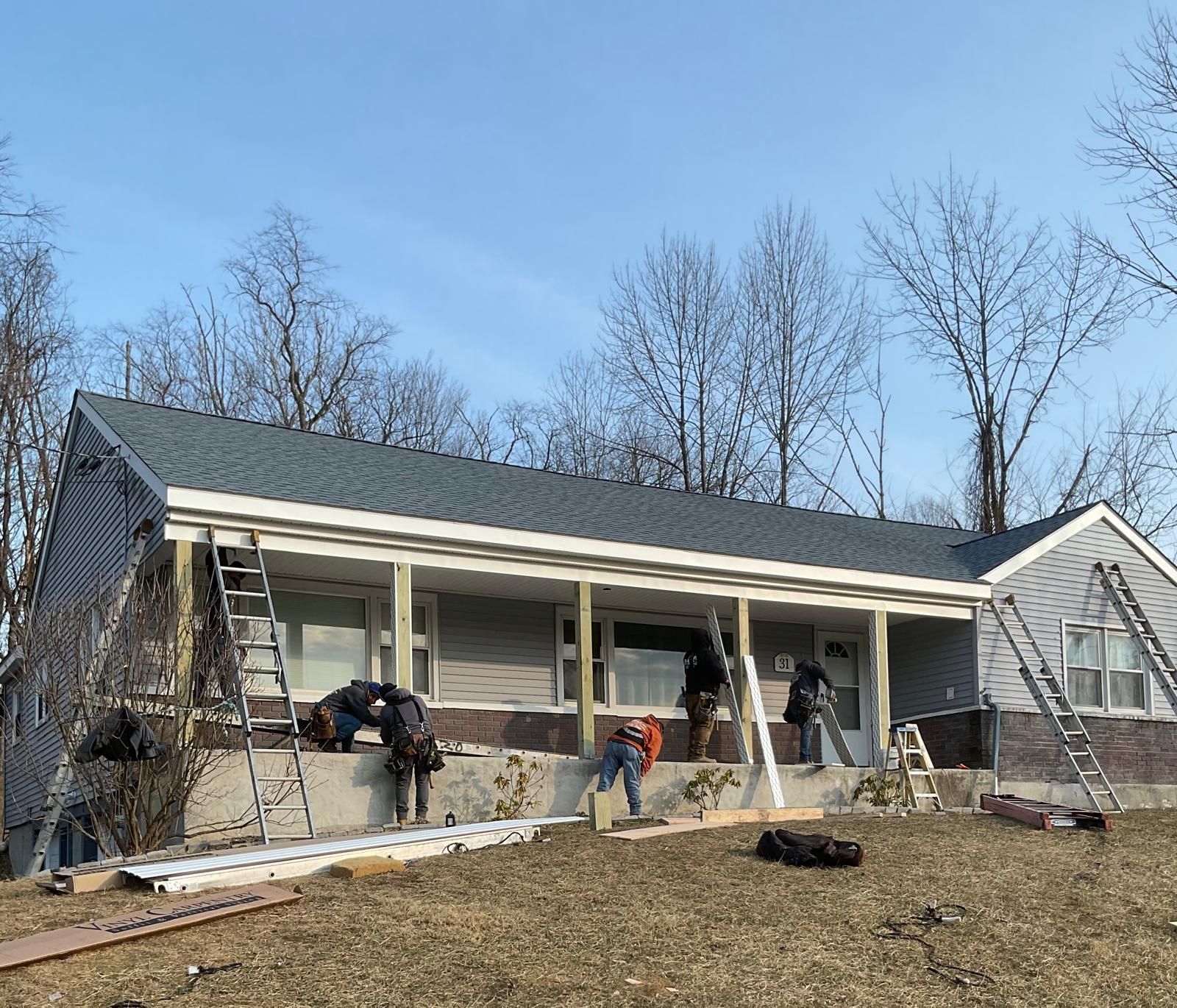 Construction workers renovate the exterior of a gray residential house with a new porch and roof under a clear blue sky.