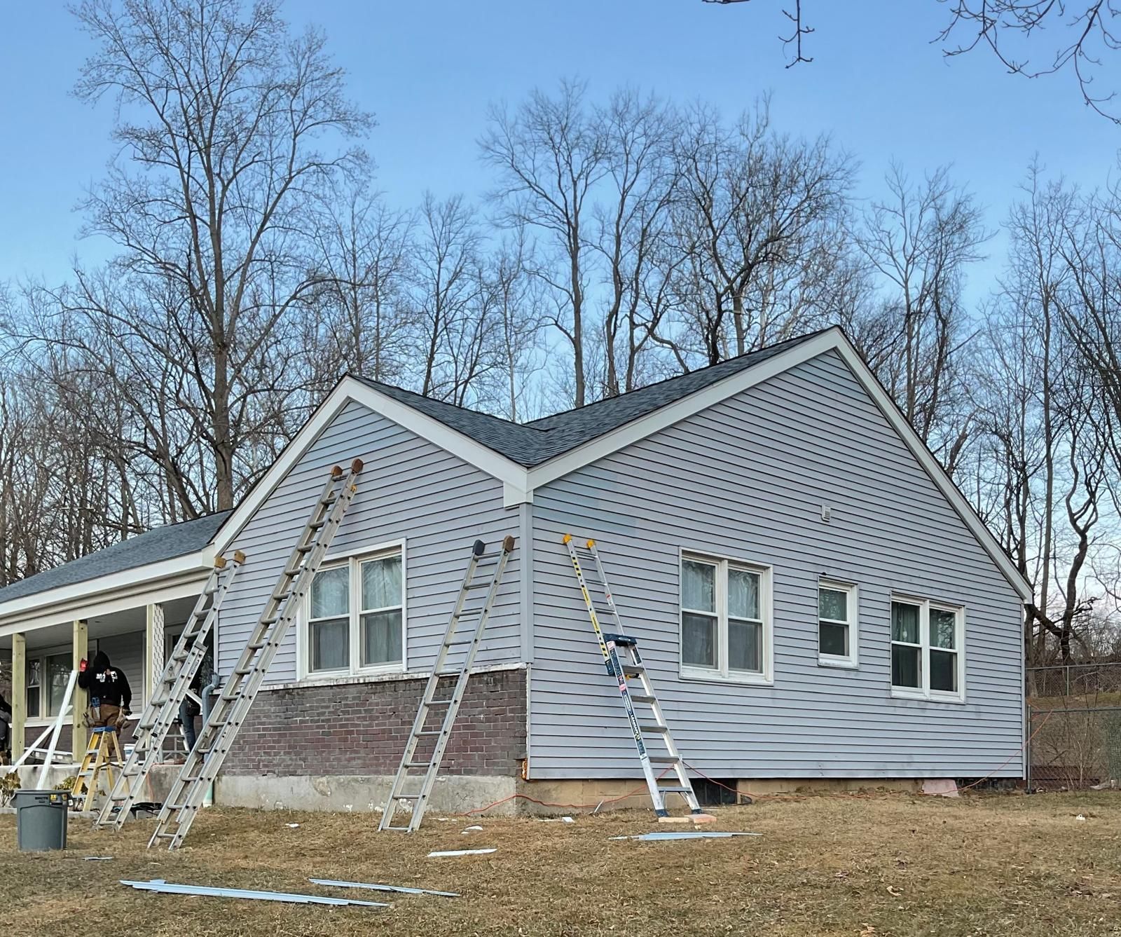 Workers using ladders to install light gray vinyl siding on the exterior of a house near a wooded area.