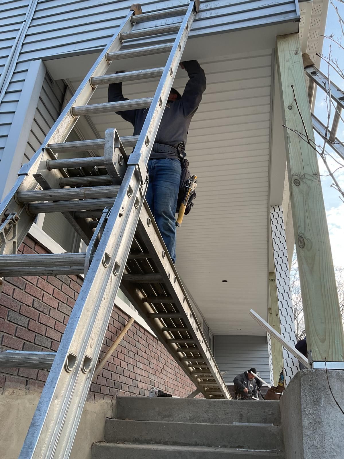 A construction worker standing on an extension ladder works on the soffit of a covered porch entrance.
