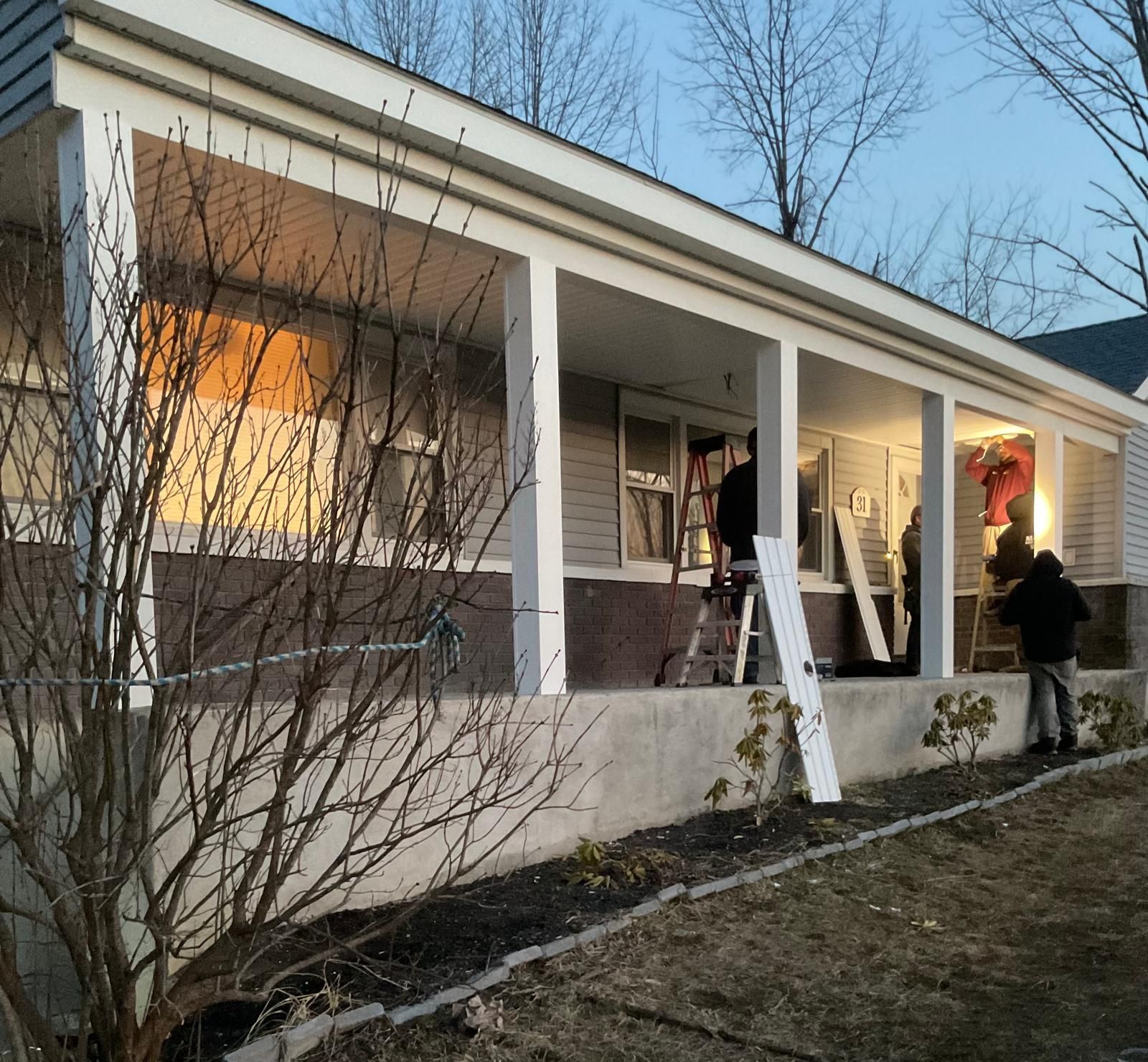 Workers in dark clothing perform renovation work on the covered front porch of a house at dusk.