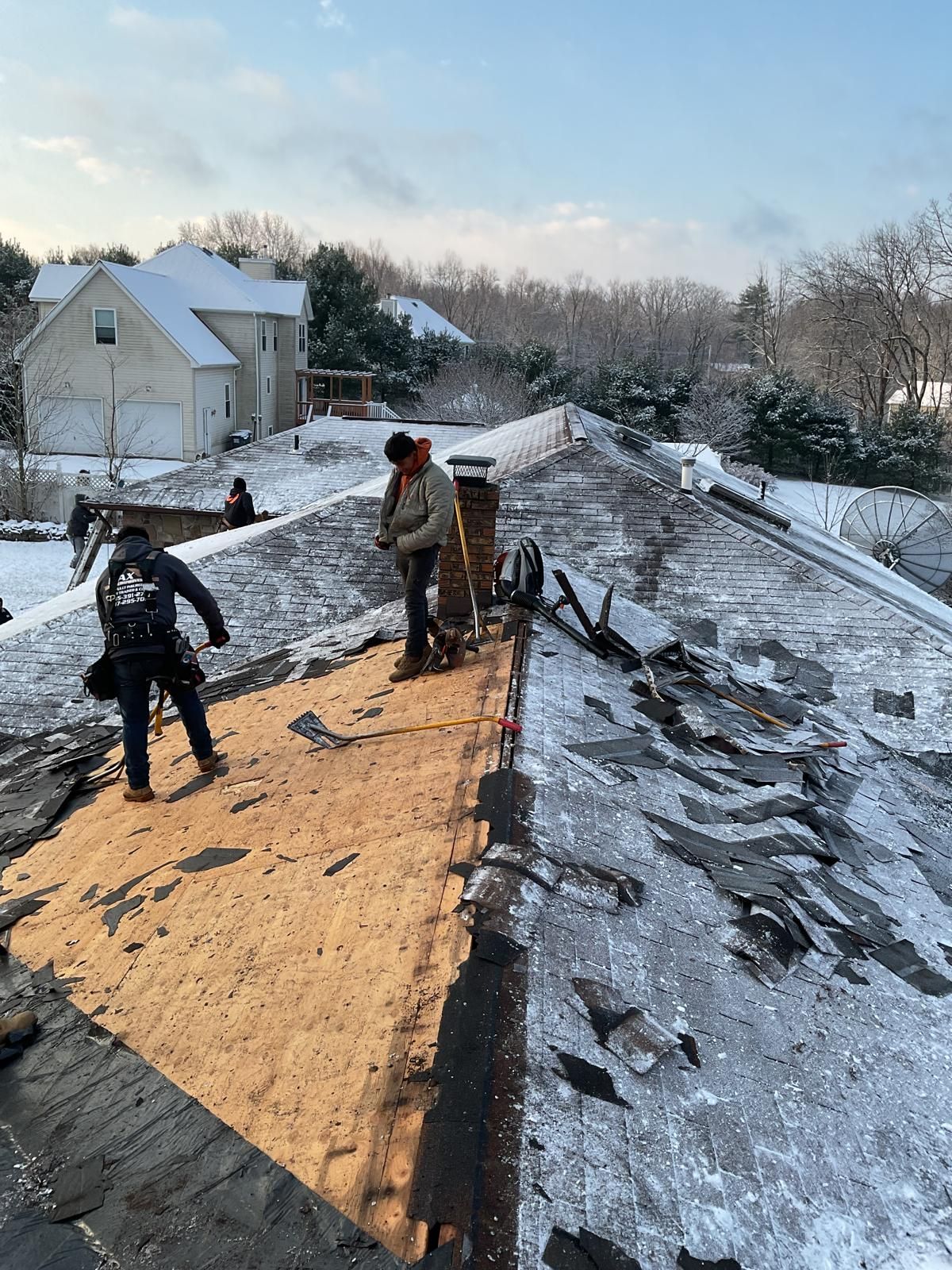 Workers remove old shingles from a snowy residential roof, exposing the wooden deck beneath.