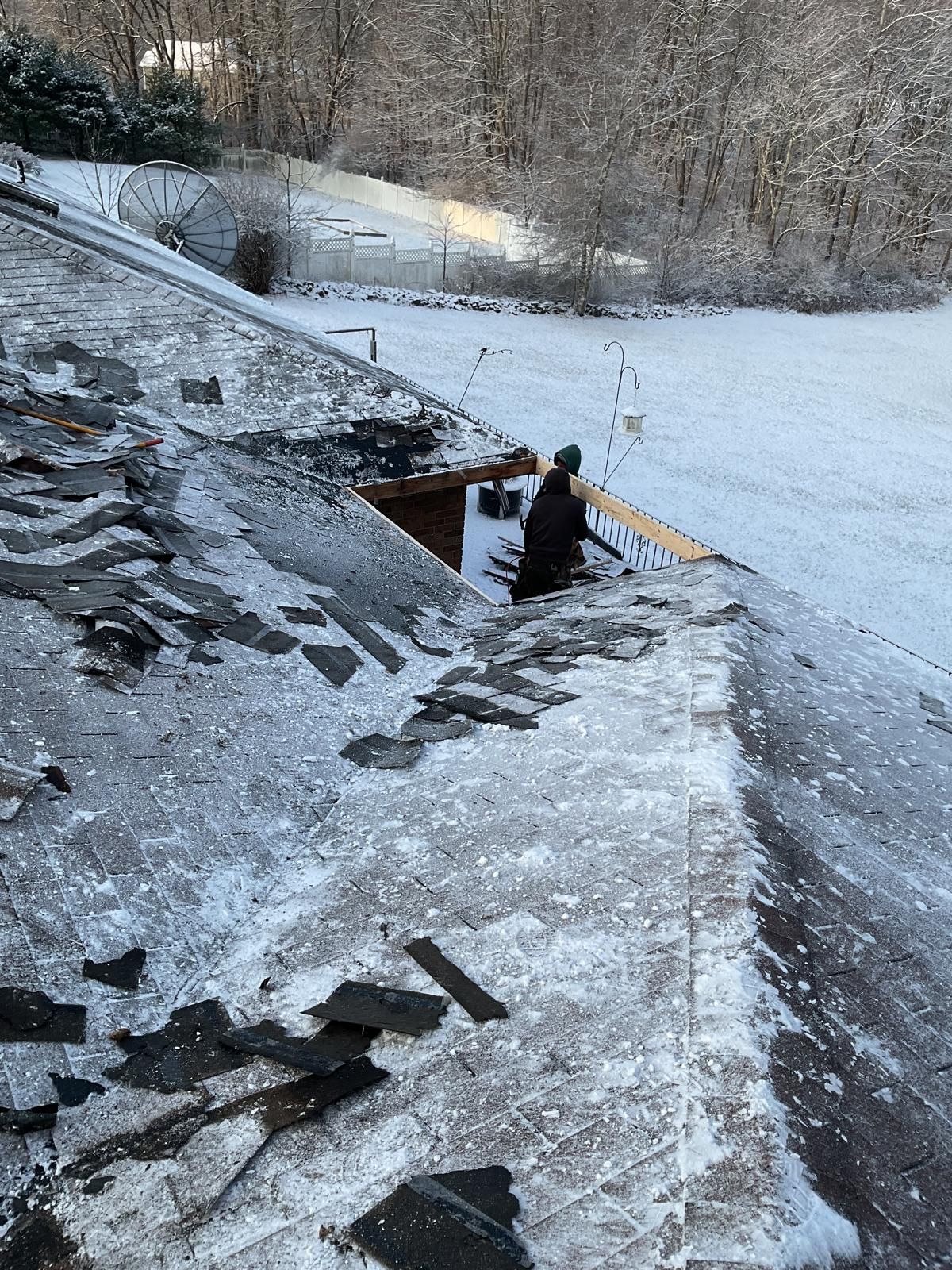 A person works on a snow-covered residential roof with shingles removed, revealing part of the underlying structure.