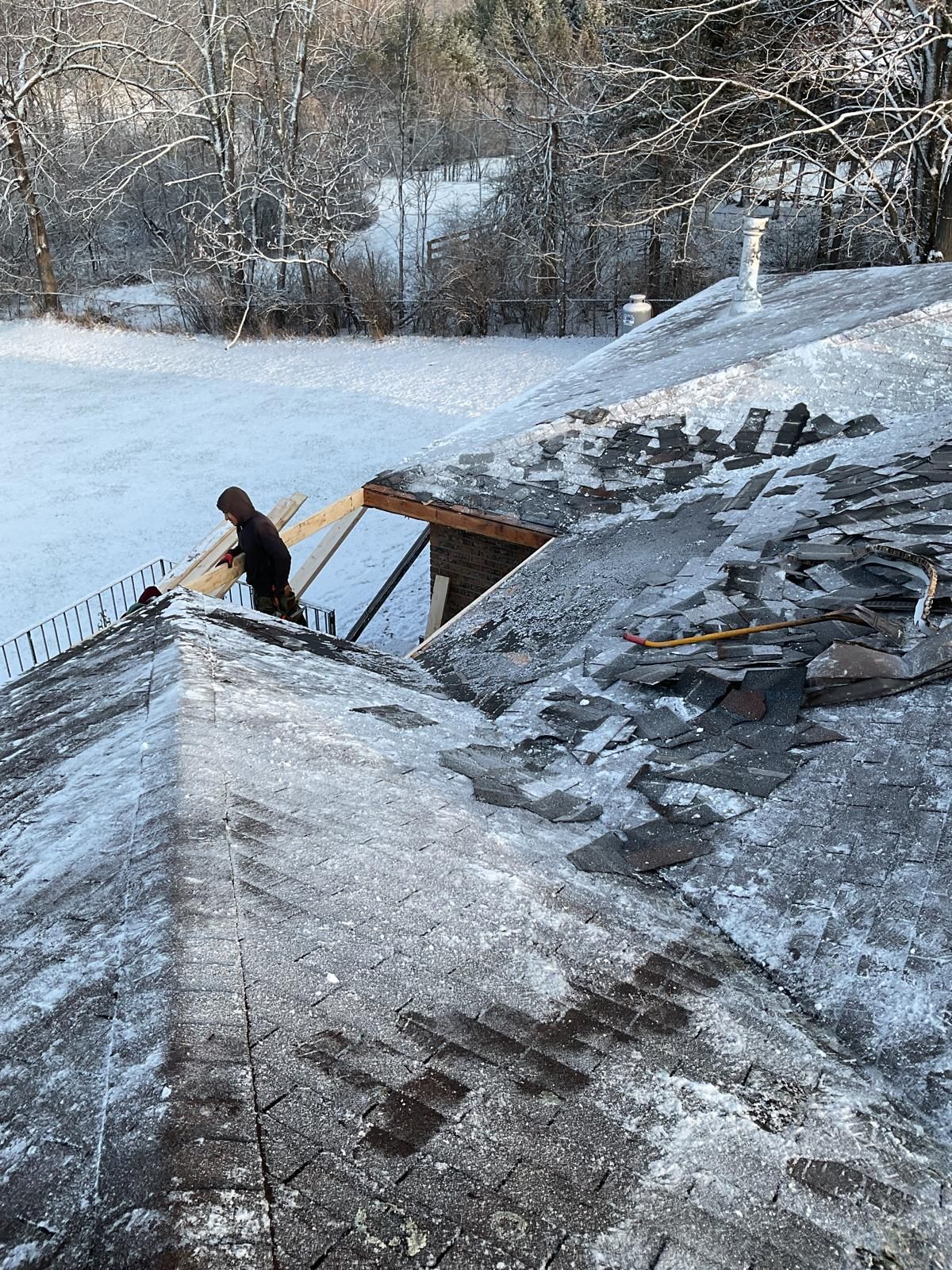 A person repairs a snow-covered residential roof, with shingles removed to expose the wooden rafters underneath.