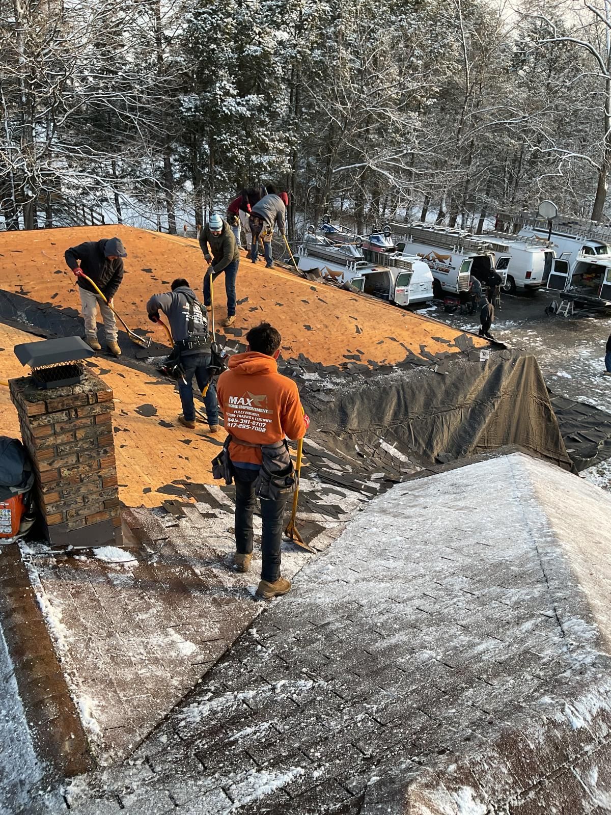A crew works on a snow-covered roof, removing old shingles to expose the wooden deck beneath on a winter day.