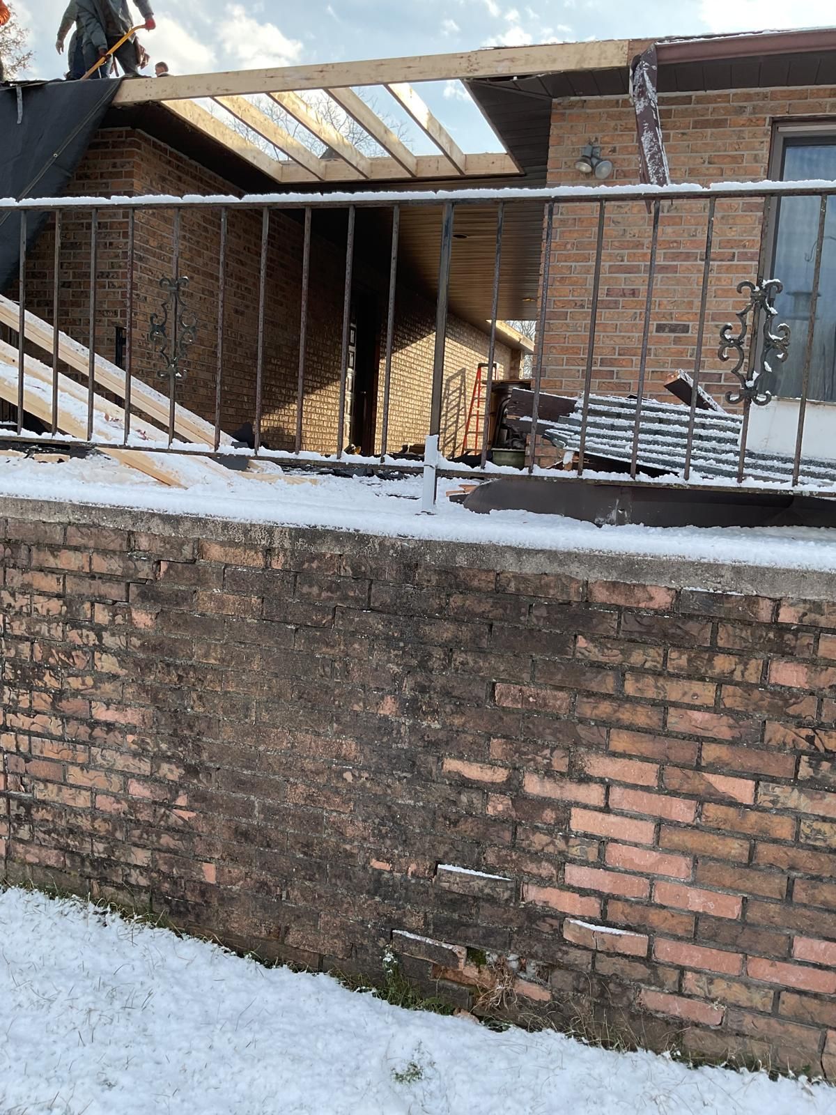 Workers perform roof repairs on a brick house during winter, with wooden joists exposed and snow on the ground.