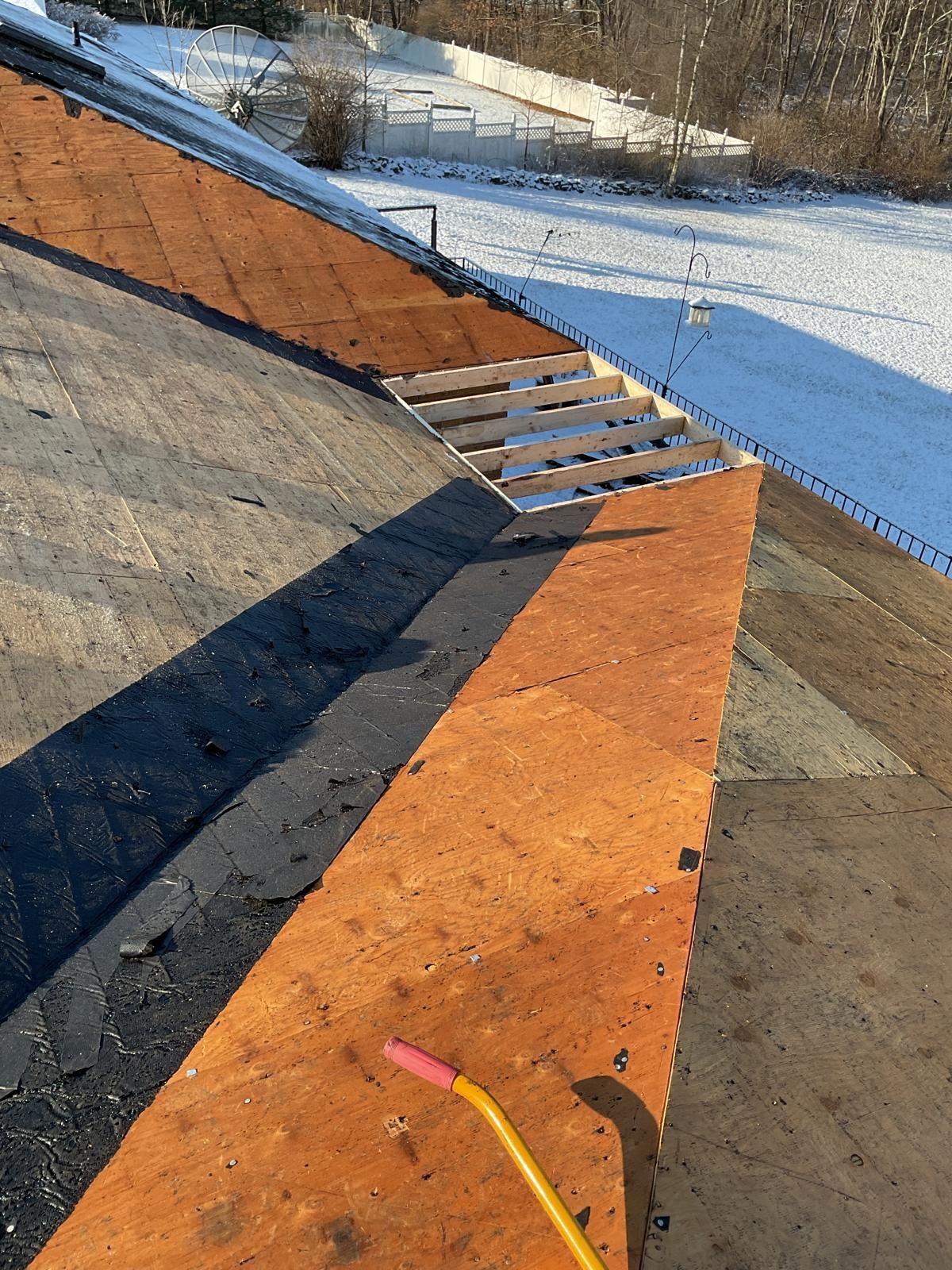 A residential roof under repair with sections of plywood sheathing and roofing underlayment exposed over a snowy yard.